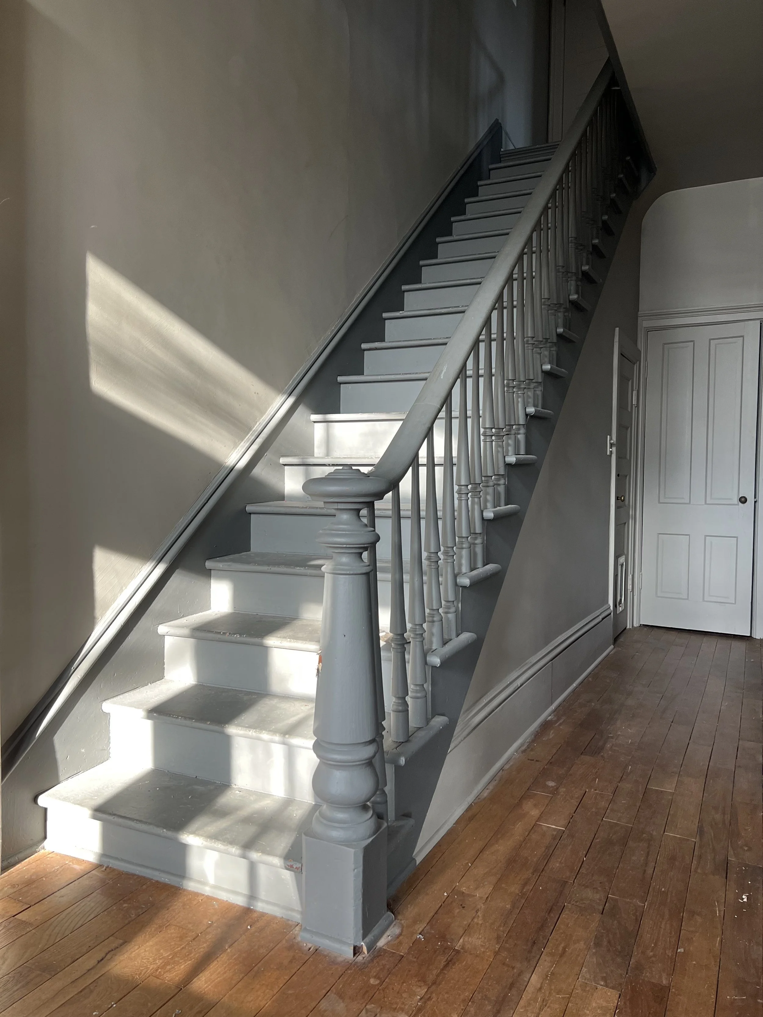 Interior of a house showing a curved gray staircase with wooden handrails and spindles, leading to an upper floor and next to a white door, on a wooden floor with sunlight streaming in.