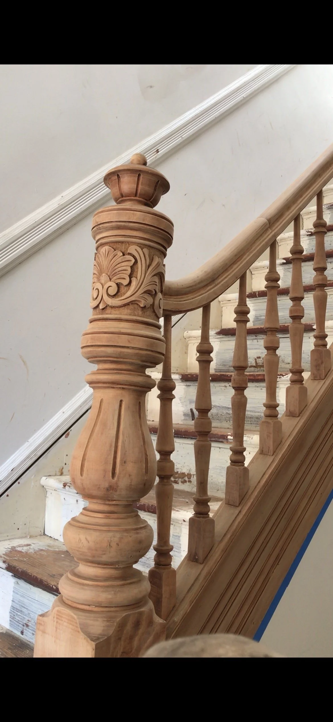 Close-up of a wooden staircase newel post with intricate carved designs, unfinished wood on stairs and trim, and a white wall with baseboard in the background.
