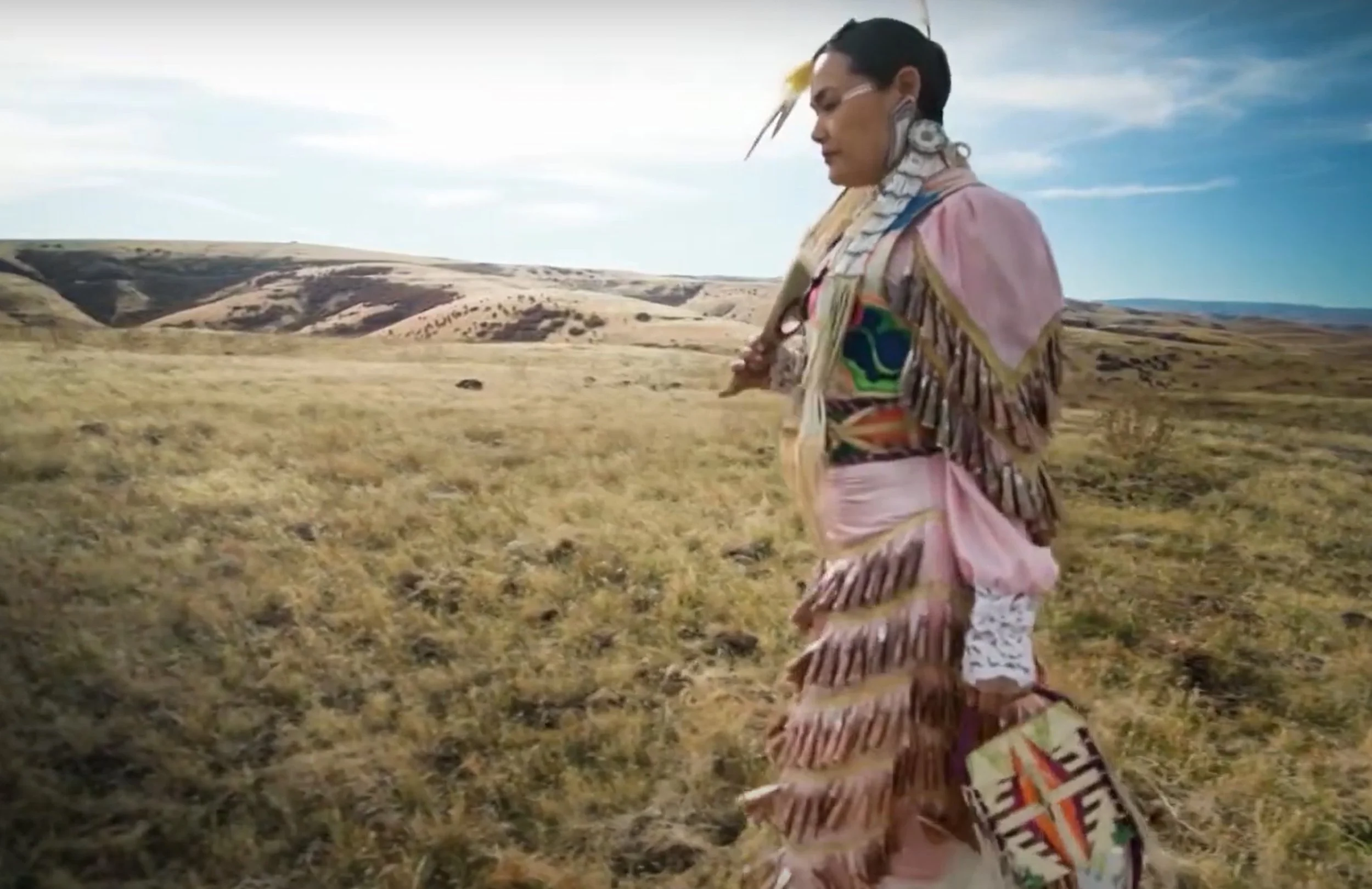 Native American woman dressed in traditional clothing walking through open grassland with rolling hills in the background.