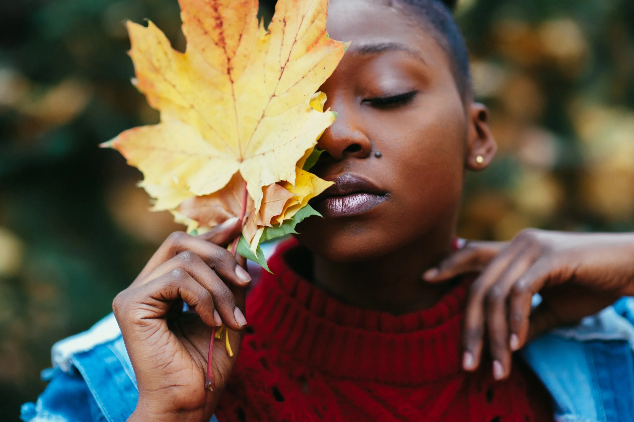 A woman holding a large yellow autumn leaf close to her face with closed eyes, wearing a red sweater and a denim jacket, with blurred fall foliage in the background.