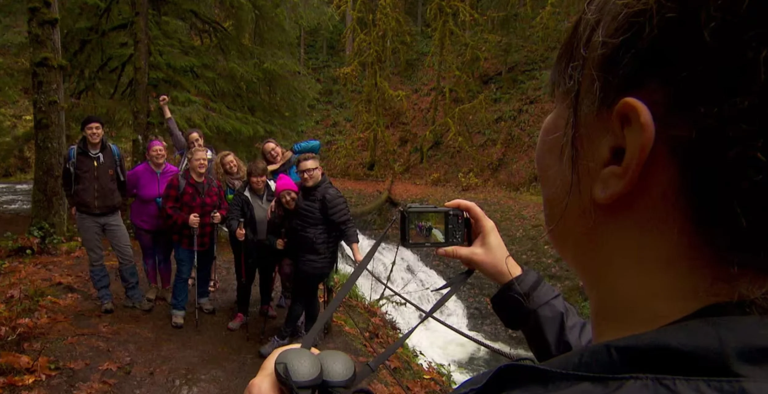 Group of people taking a photo in a forested area near a waterfall, surrounded by trees with autumn leaves.