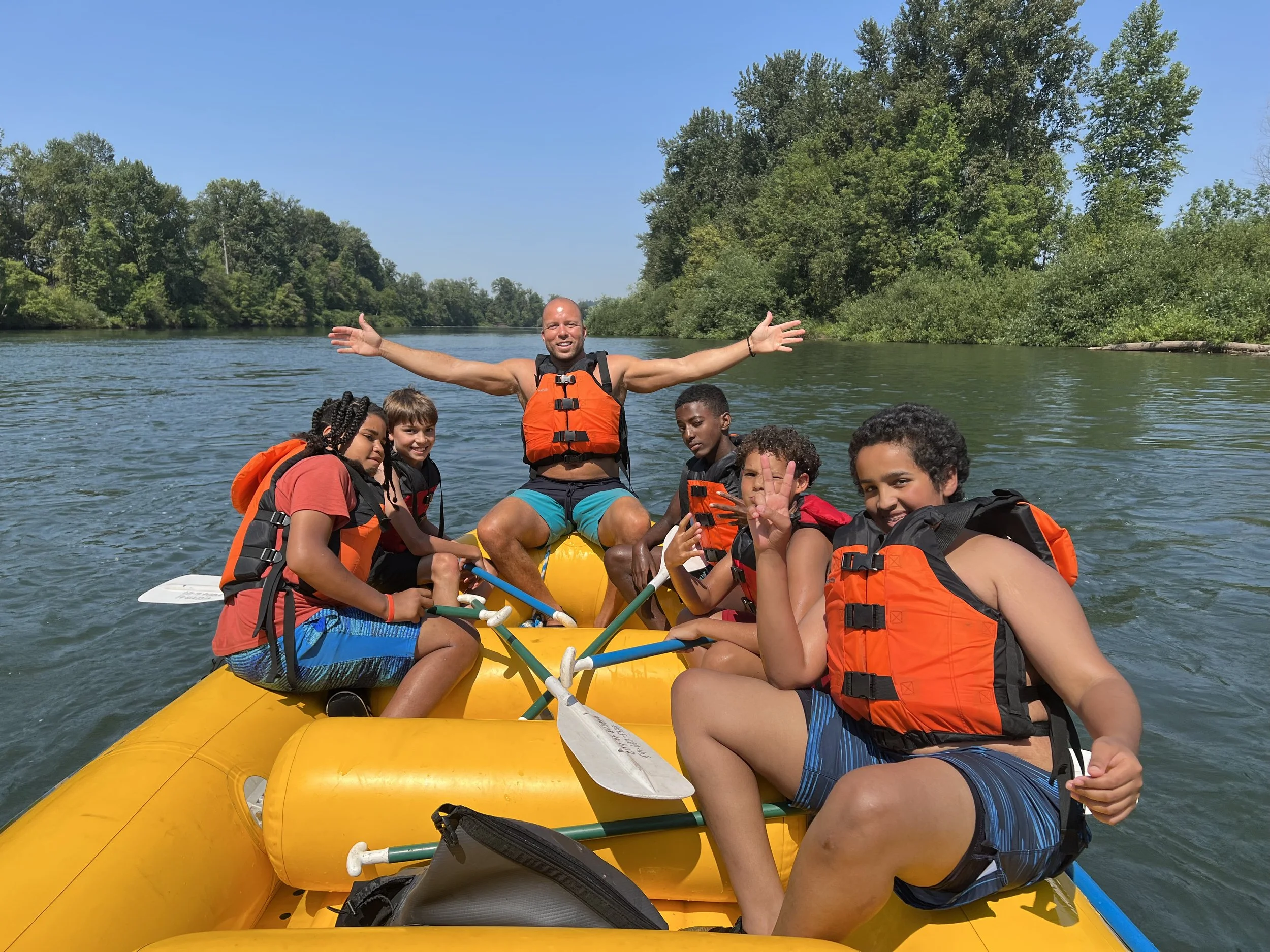 Group of teens and an adult (Founder/Executive Director and Outdoor Leader Niles Mittasch) on a yellow inflatable raft in the water, with trees and blue sky in the background, all wearing life jackets.
