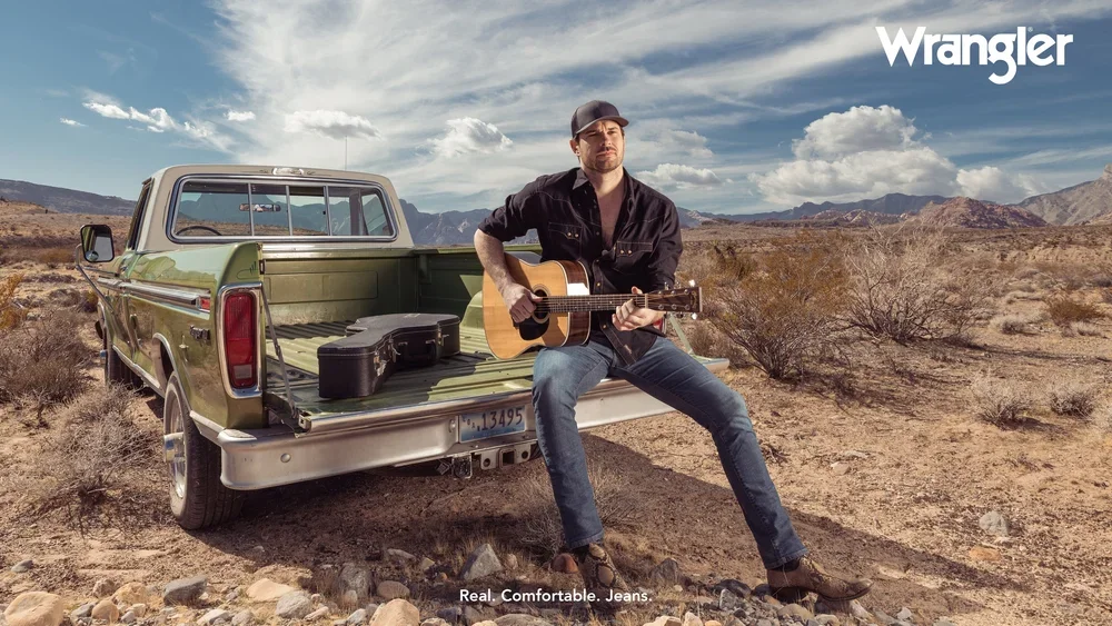 Man holding a guitar beside a pickup truck in a desert commercial image with Wrangler branding.