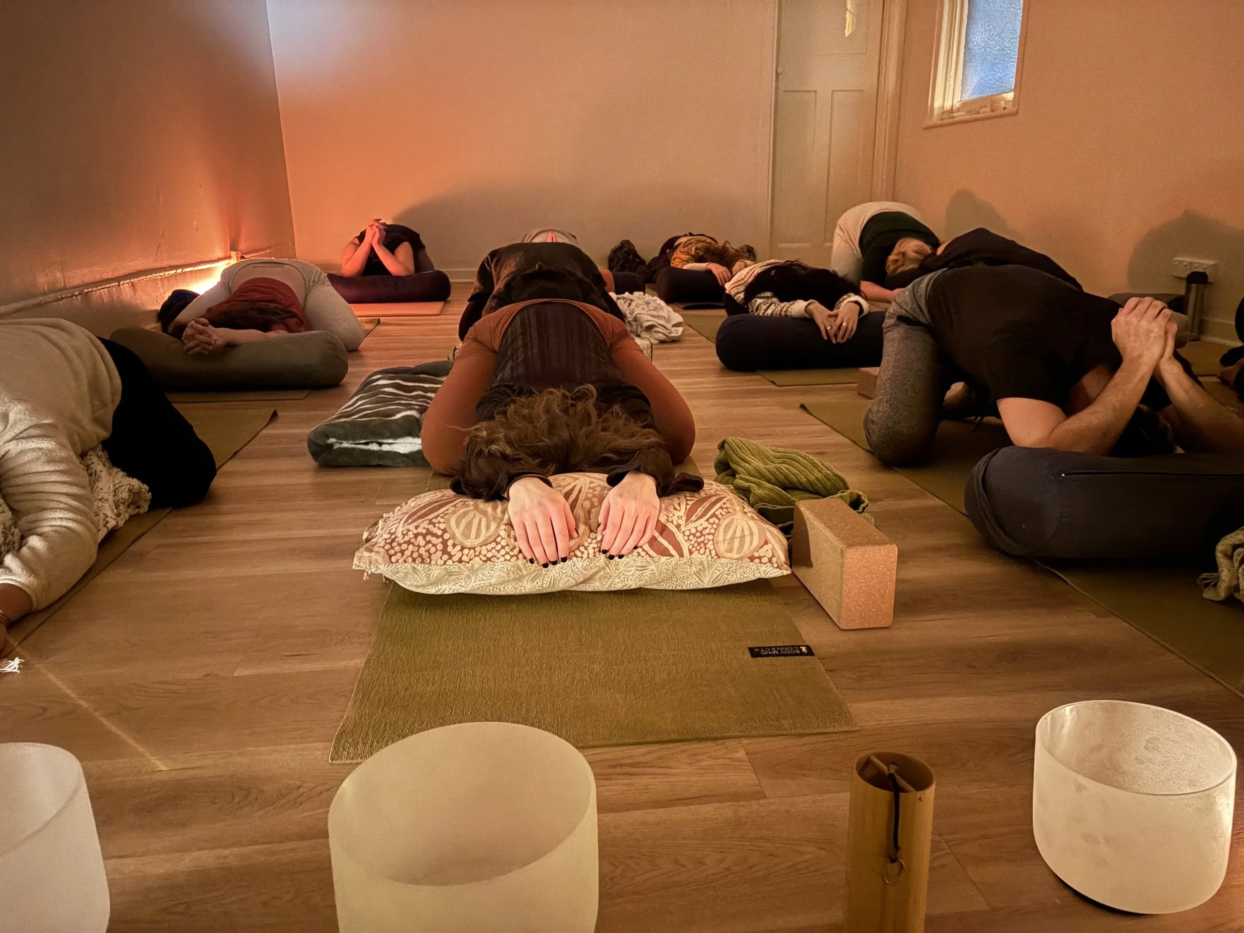 People practicing yoga in a dimly lit room with orange lighting, lying on yoga mats with hands clasped behind their heads in a stretching pose.