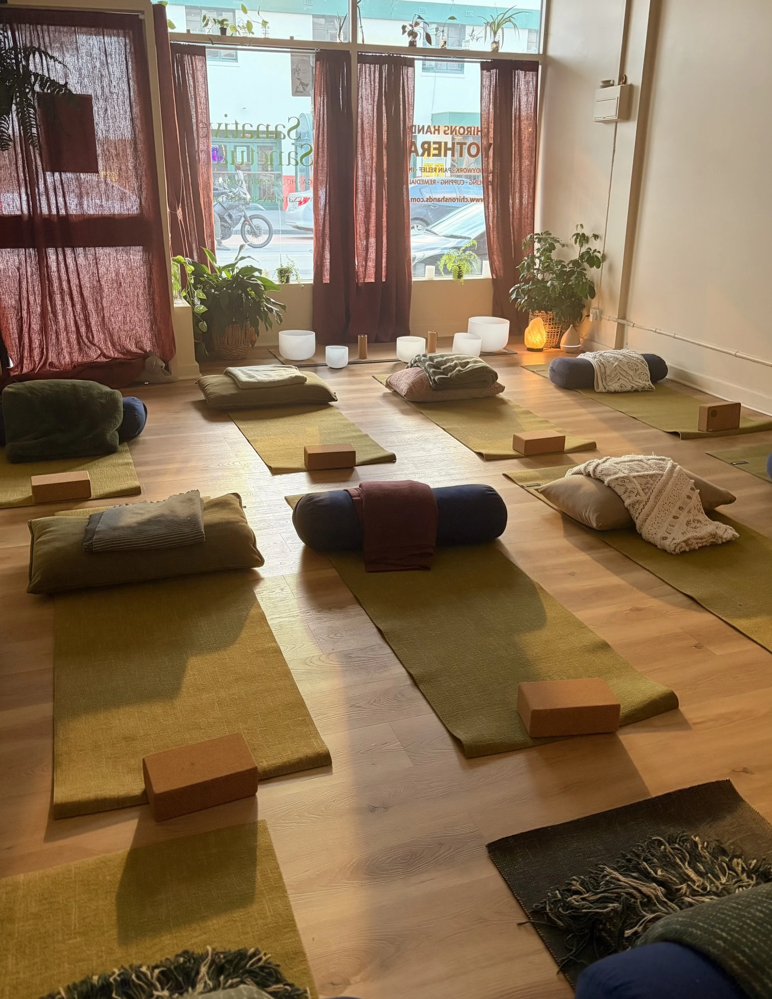 A yoga or meditation studio with mats, pillows, and blankets arranged on a wooden floor. Large windows with plants and curtains let in natural light outside.