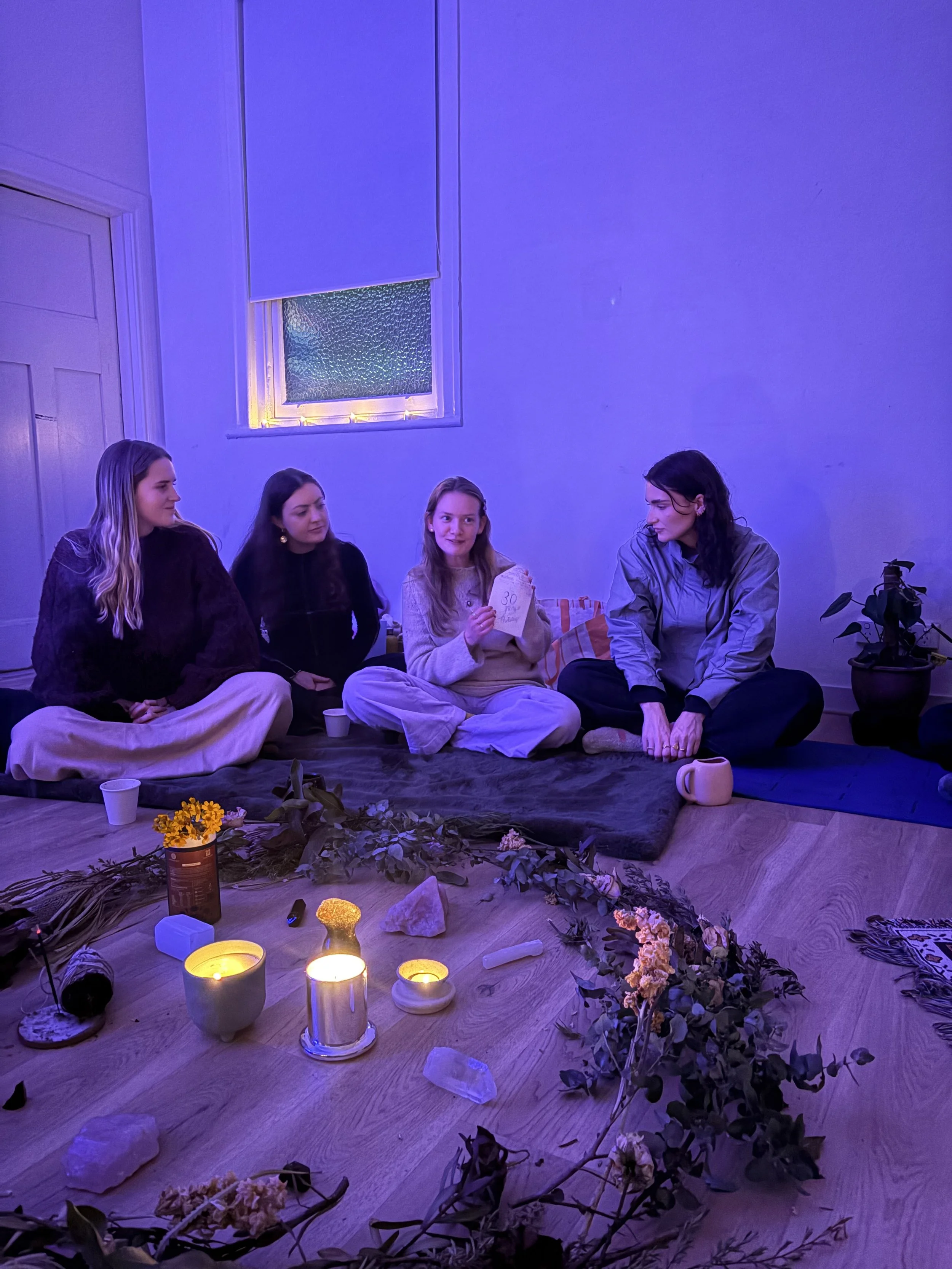 Four women sitting on the floor in a softly lit room, surrounded by candles and flowers, engaging in a group activity or discussion.