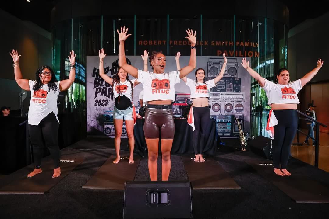 A group of five women practicing yoga on a stage during a fitness event, all wearing white T-shirts with a red camera graphic and the text 'FITDC'.