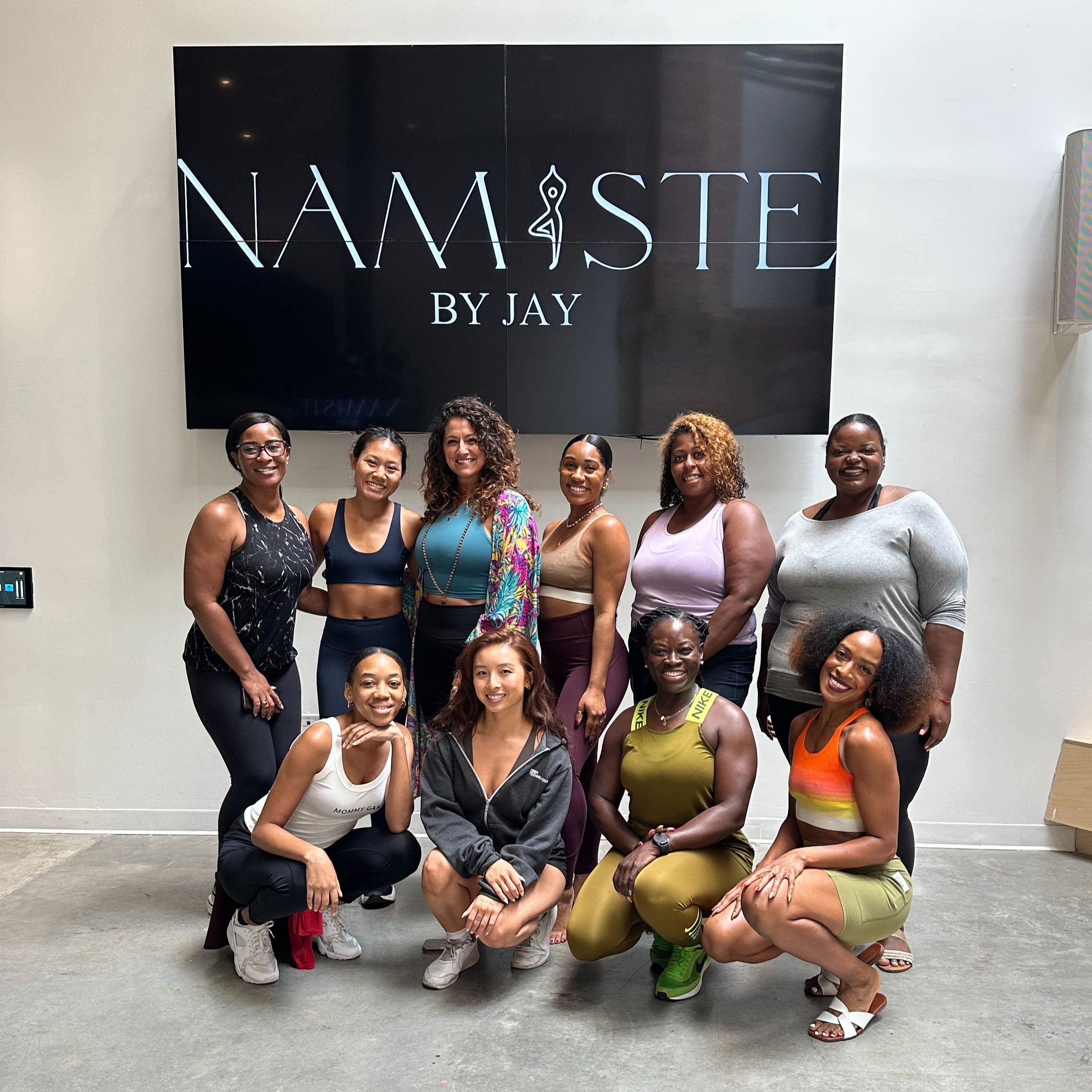 Group of eleven women in workout clothing posing in front of a large black screen that reads 'Namaste by Jay' in a fitness studio.