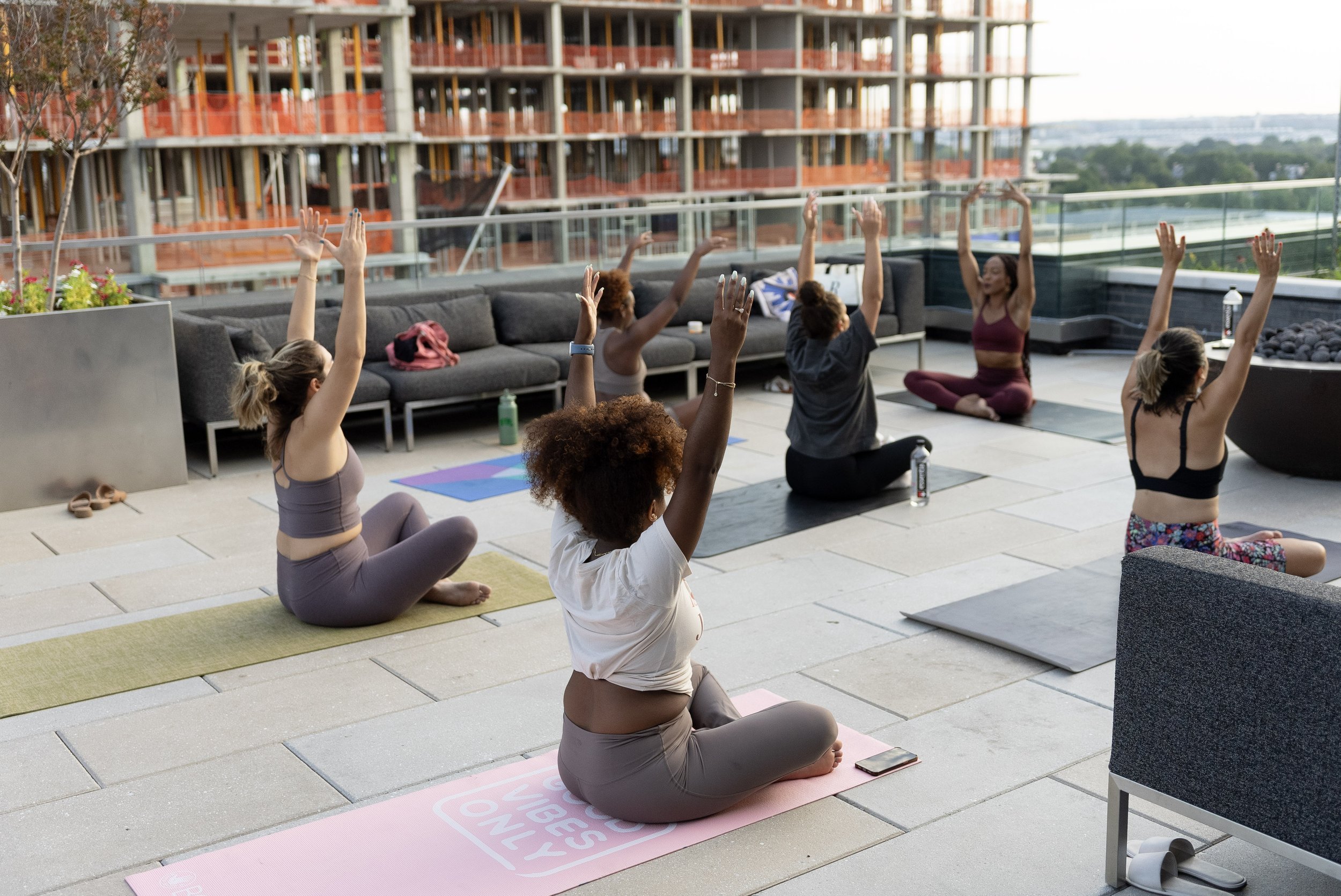 Six women participating in a rooftop outdoor yoga class, sitting on yoga mats with arms raised, facing an instructor.