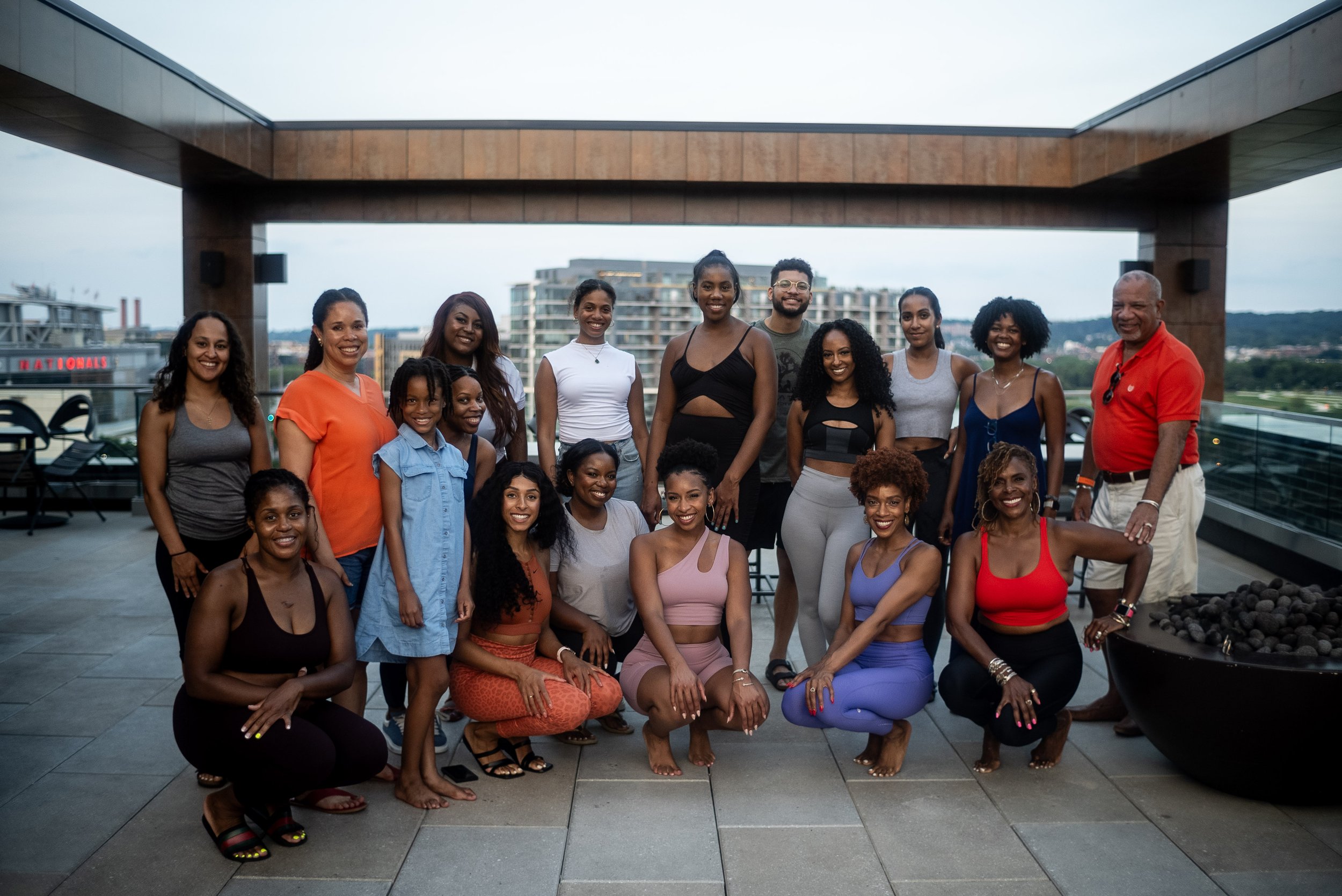 Group of diverse women and one man gathering on a rooftop balcony with cityscape in the background, some wearing activewear, smiling for the photo.