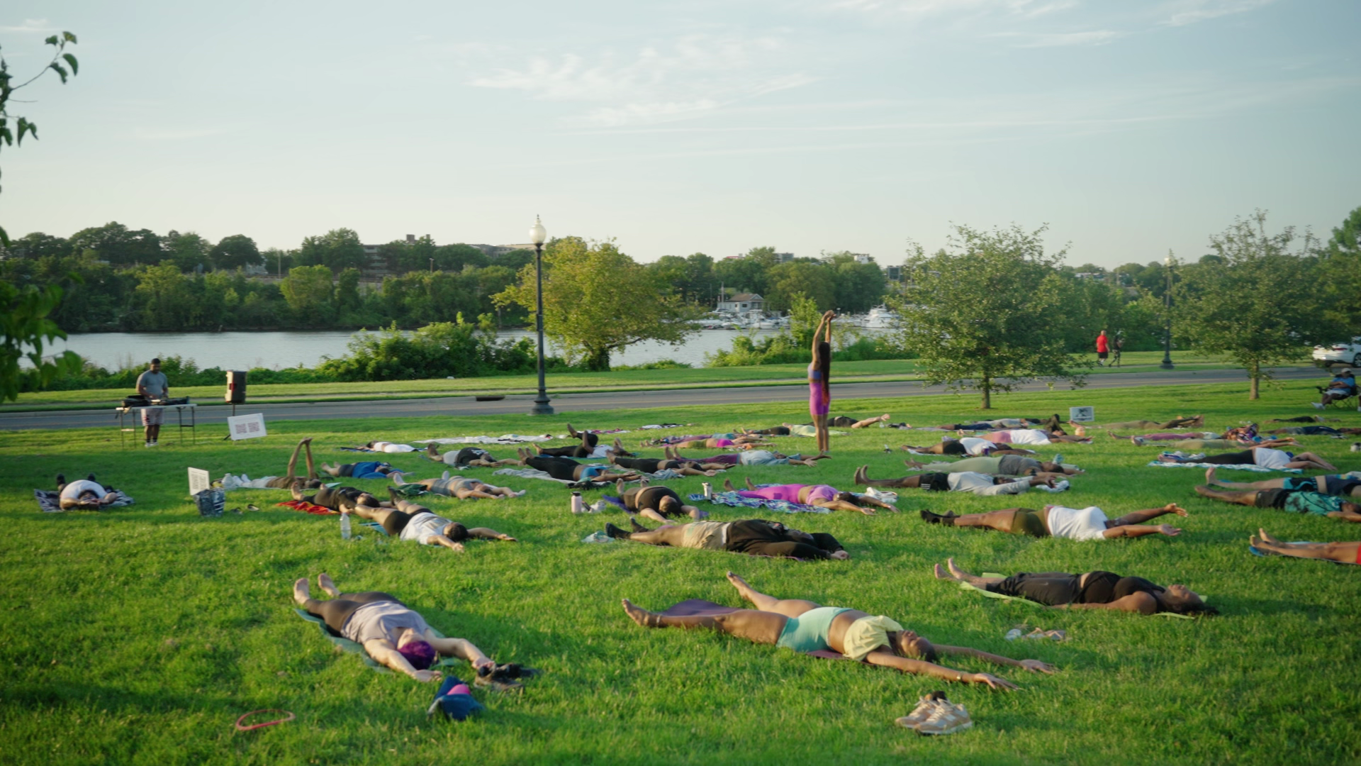 Group of people practicing outdoor yoga on grass near a river, with trees and a walking path in the background.