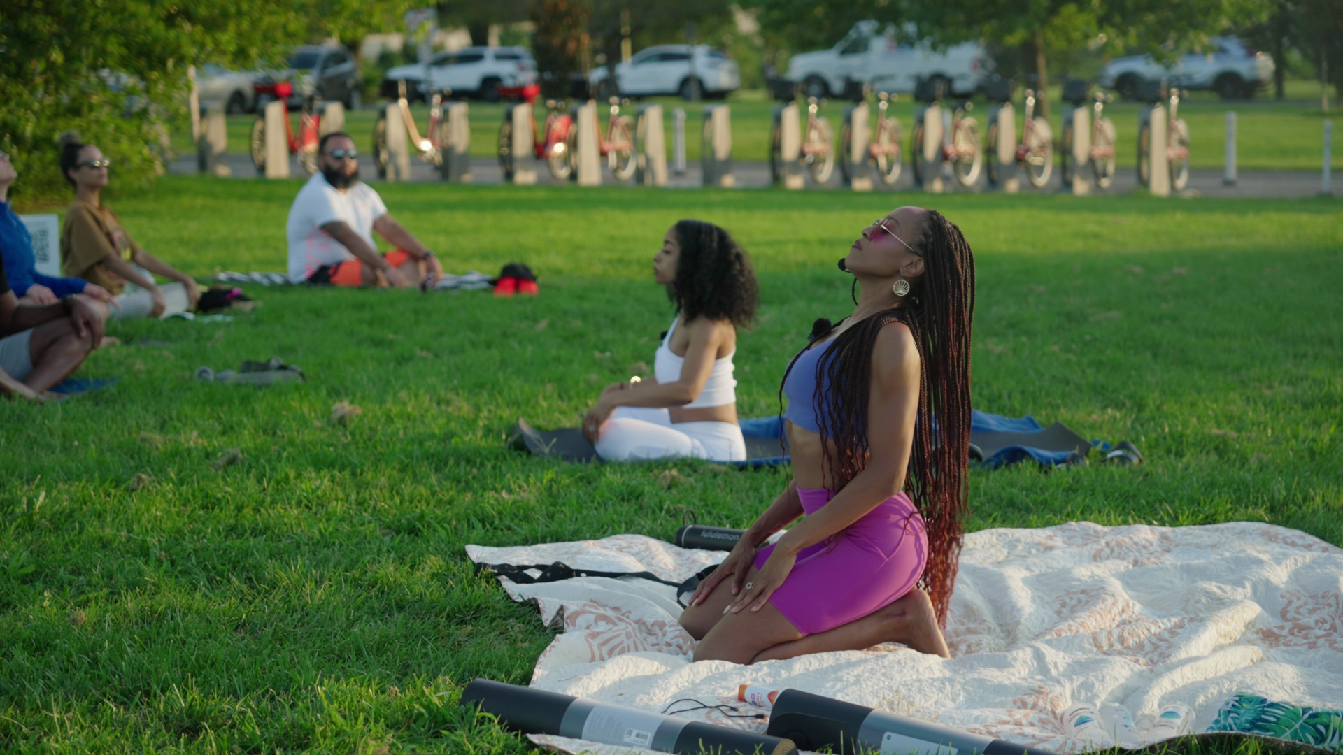 A group of people practicing yoga outdoors on a grassy field, sitting on mats and blankets, with bicycles and parked cars in the background.