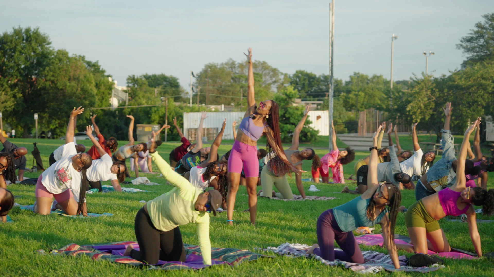 Group of people participating in an outdoor yoga class on a grassy field, performing a side stretch pose under clear sky, surrounded by trees and urban structures.