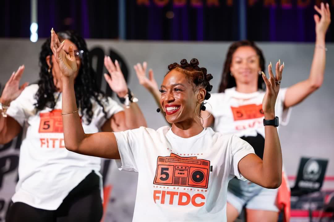 A group of women dancing and smiling at a fitness event, wearing matching white T-shirts with an orange graphic of a boombox and the text "5th Edition FITDC".