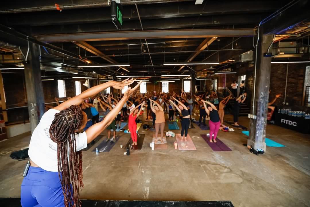 A large indoor yoga class with many participants standing on yoga mats, stretching with arms overhead, led by an instructor at the front. The room has industrial-style decor with exposed beams and brick walls.