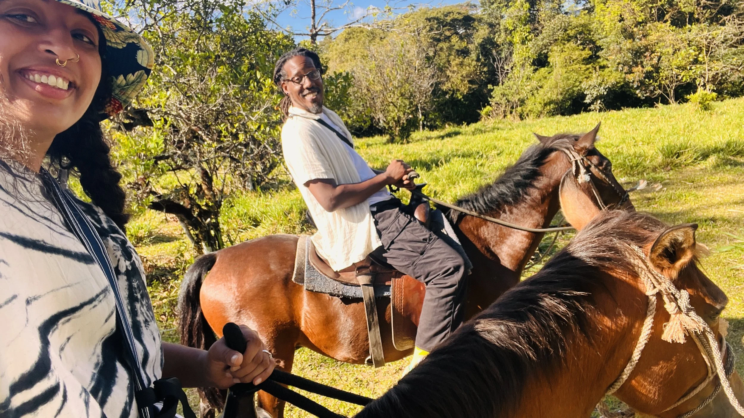 Two women and a man riding horses on a sunny day in a grassy, wooded area.