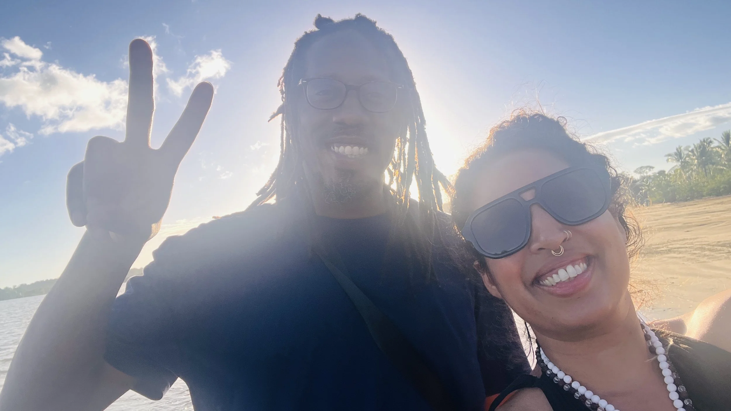 A man and woman smiling at the camera, taking a selfie outdoors during daytime with a clear sky, some clouds, and a sandy beach with trees in the background. The man is making a peace sign with his hand. Both are wearing sunglasses.