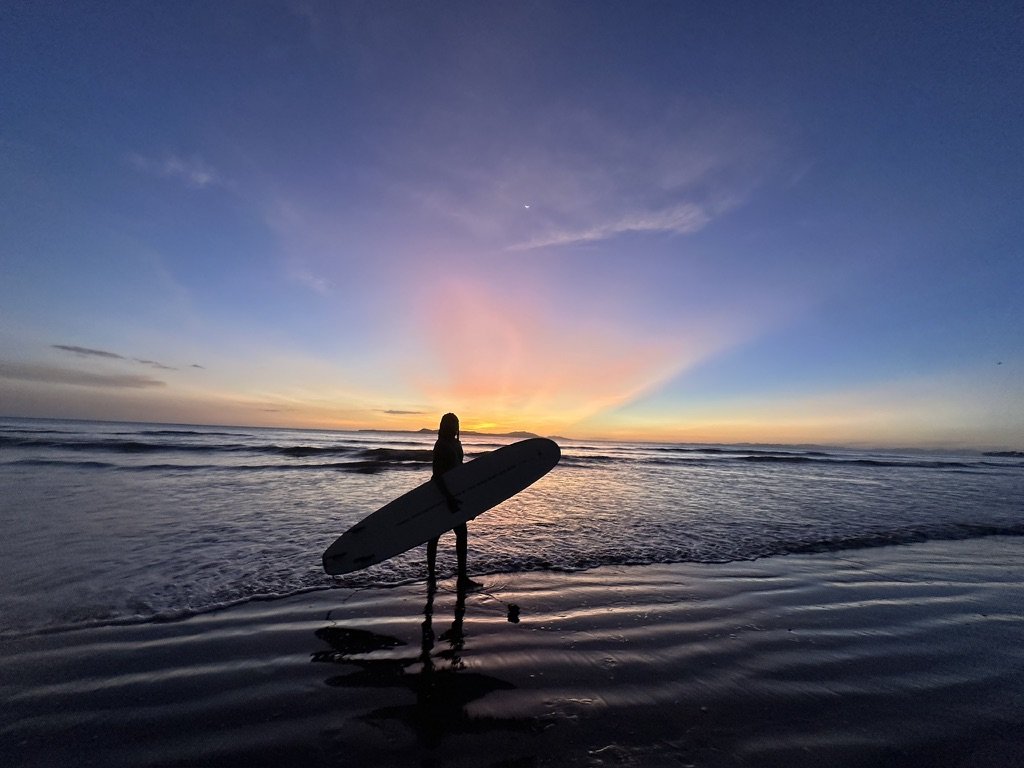 Silhouette of a person holding a surfboard on a beach during sunset, with colorful sky and ocean waves.