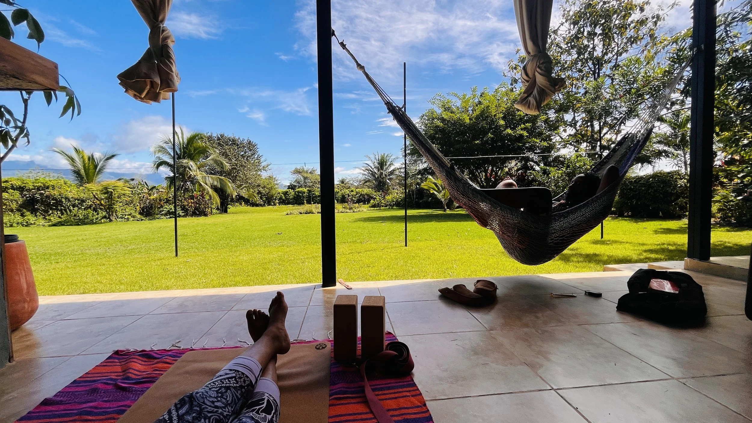 Person relaxing on a yoga mat and person lying in a hammock outdoors on a sunny day with green grass and trees.