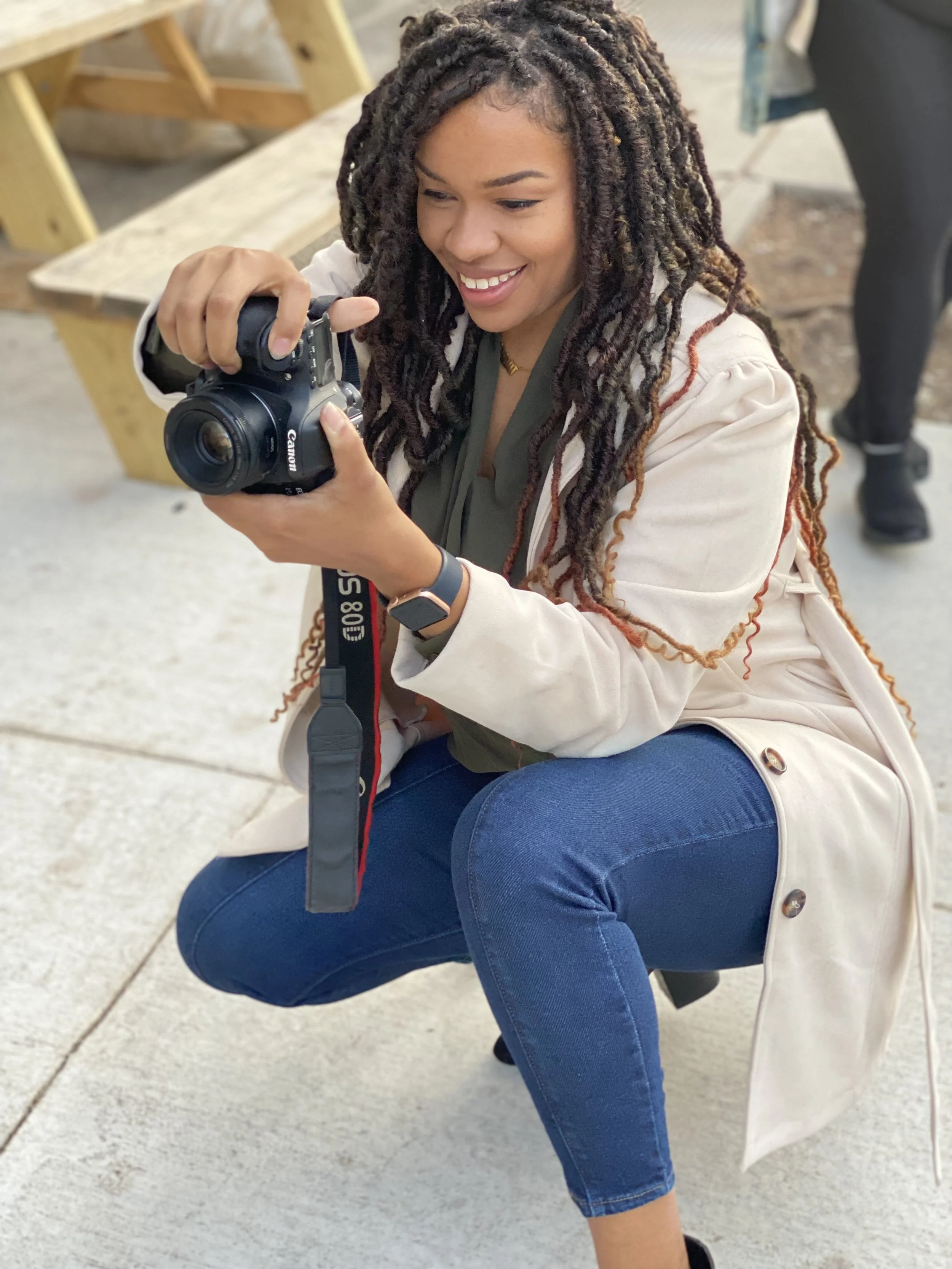 A woman with curly dreadlocks smiling and looking at a DSLR camera she is holding, crouched on one knee outdoors.
