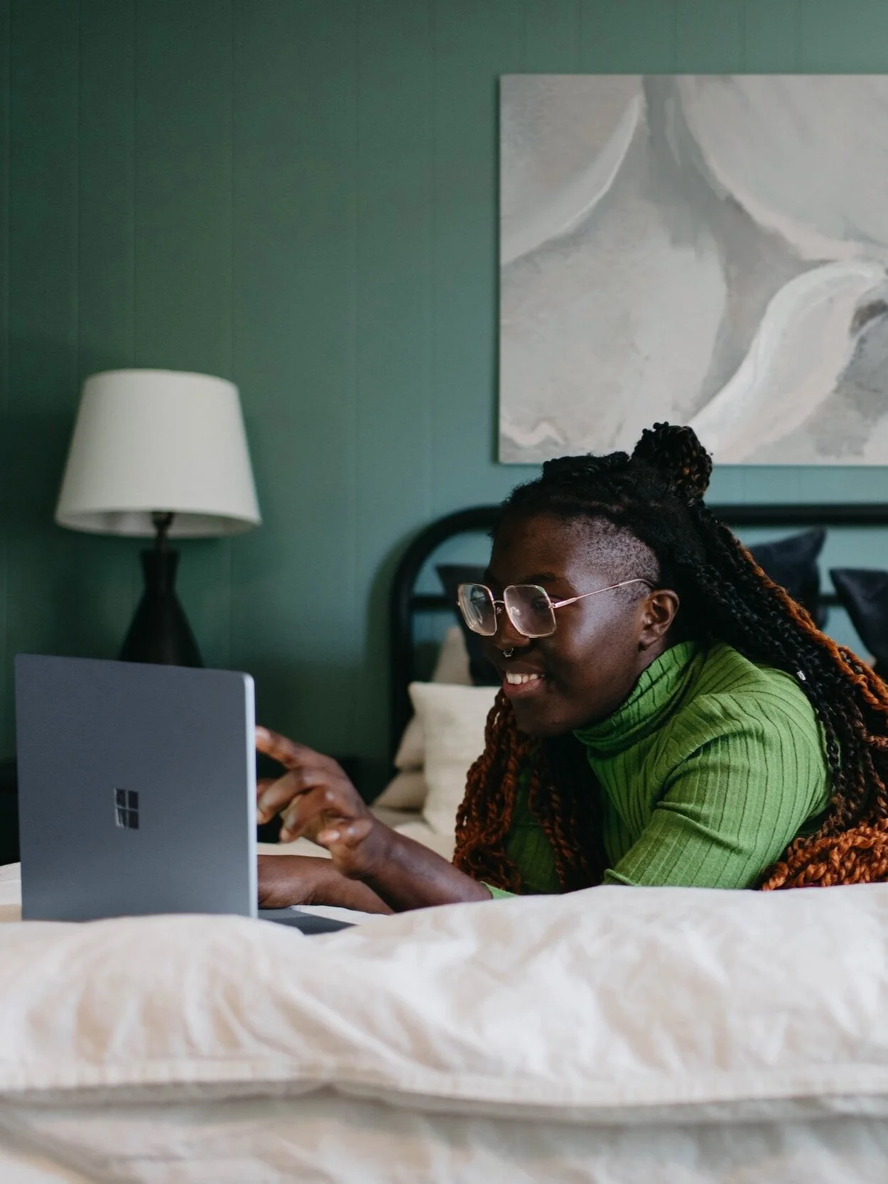 Person with glasses and braids using a tablet in bed with green walls, a white lamp, and abstract art on the wall.