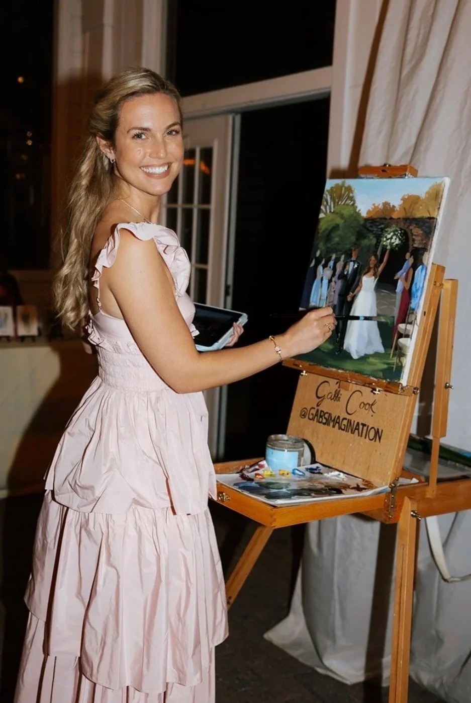 A woman in a pink dress smiling and painting at a wedding reception. The painting depicts a wedding scene outdoors, with a bride and groom and guests around them.