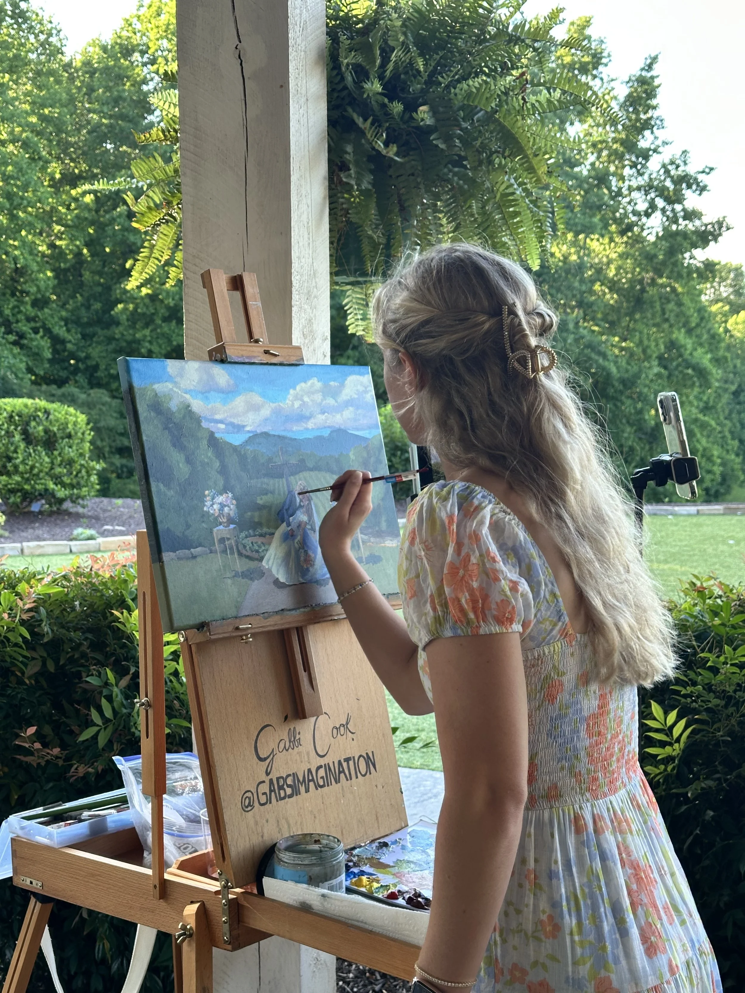 A young girl with wavy blonde hair, wearing a floral dress, is painting outdoors on an easel. She is holding a paintbrush and working on a landscape painting, with greenery and trees in the background.