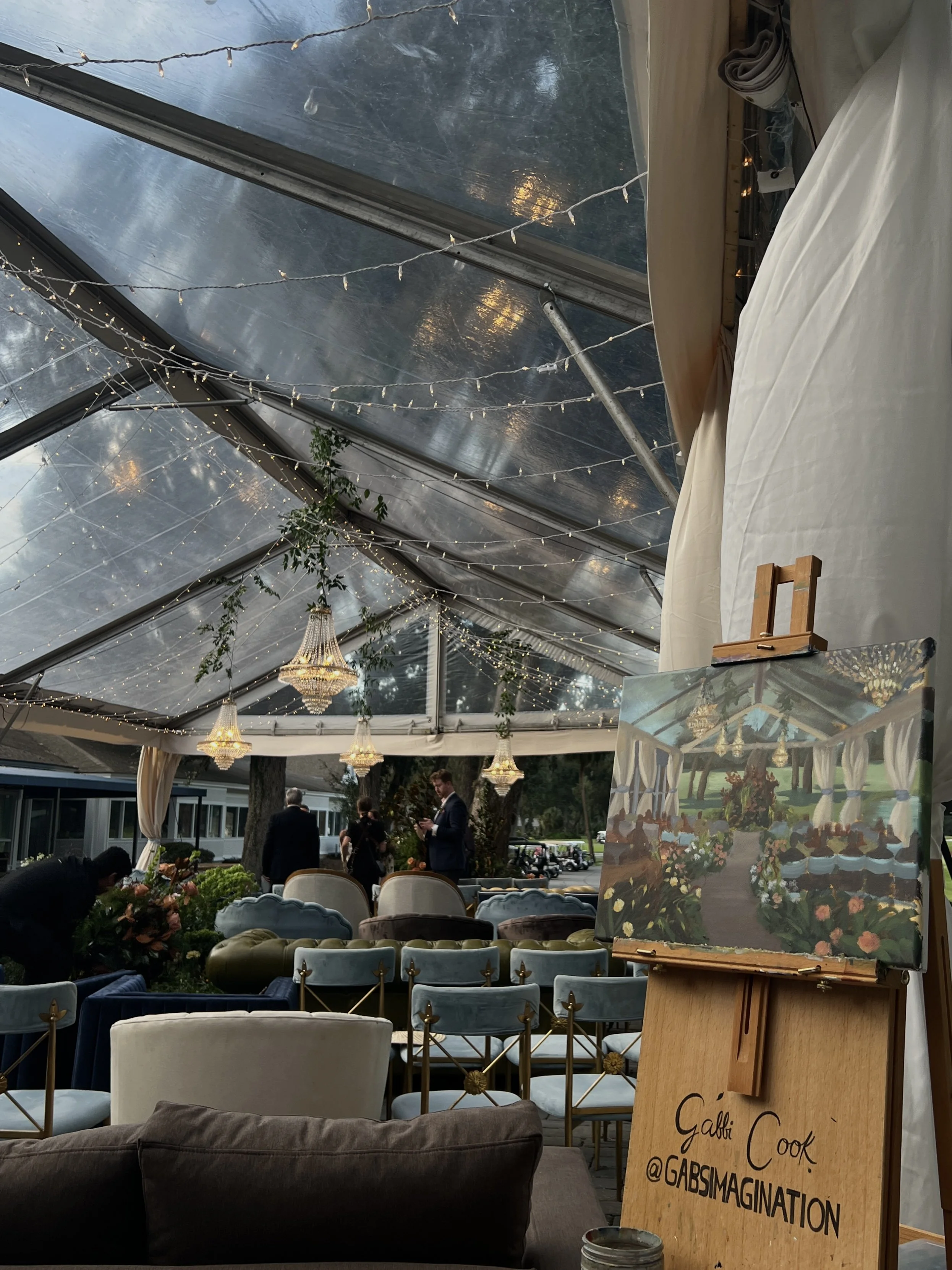 Inside a decorated tent at an event, with string lights and chandeliers hanging from the ceiling, chairs arranged for guests, and a small painting on an easel with the artist's name and social media handle.