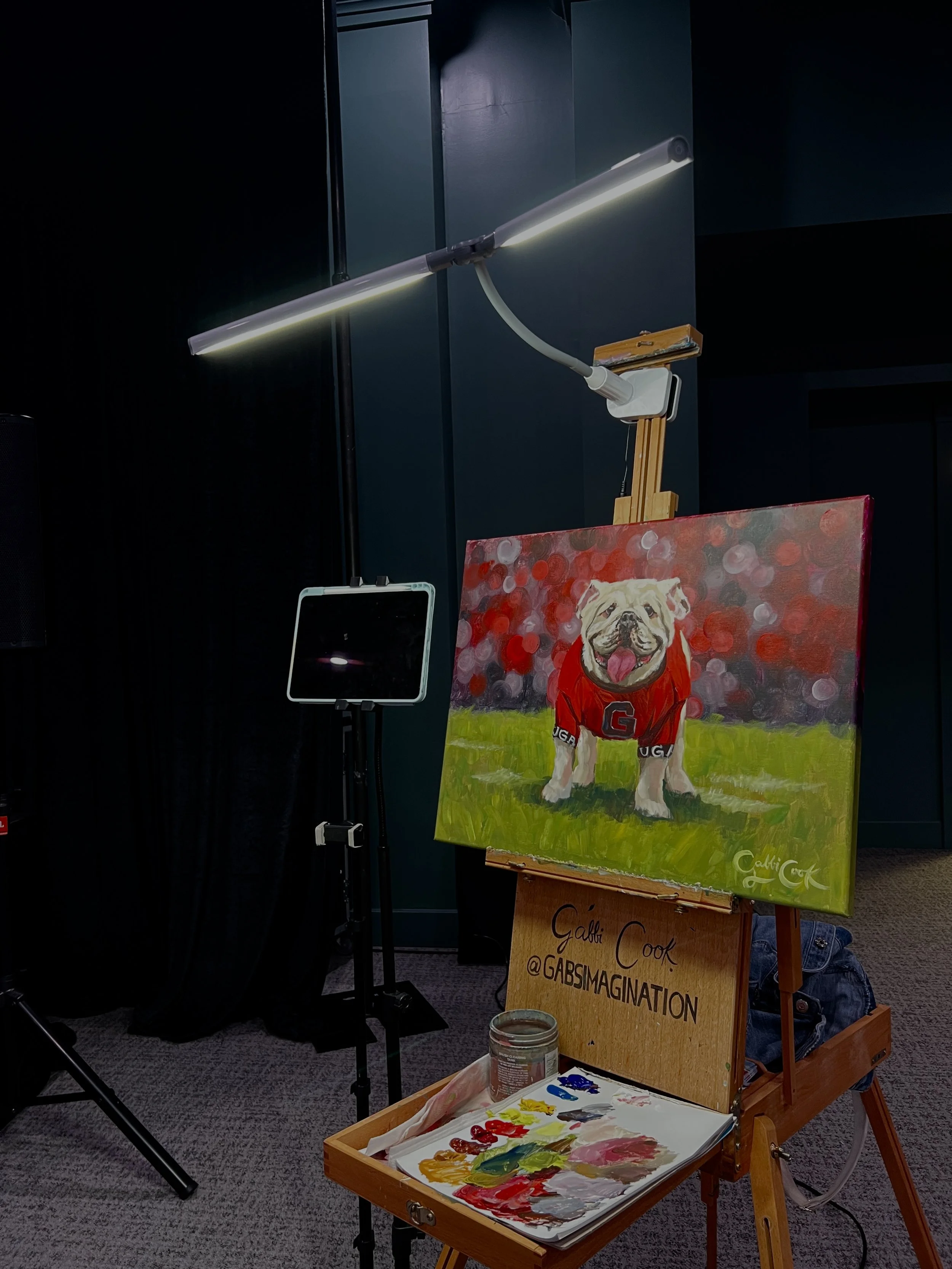 Painting of a bulldog wearing a red sports jersey, standing on grass with a blurred red background, on an easel with a sign reading 'Gabbi Cook @GABSMAGINATION'. An artist's palette with paint and brushes is on a small table.