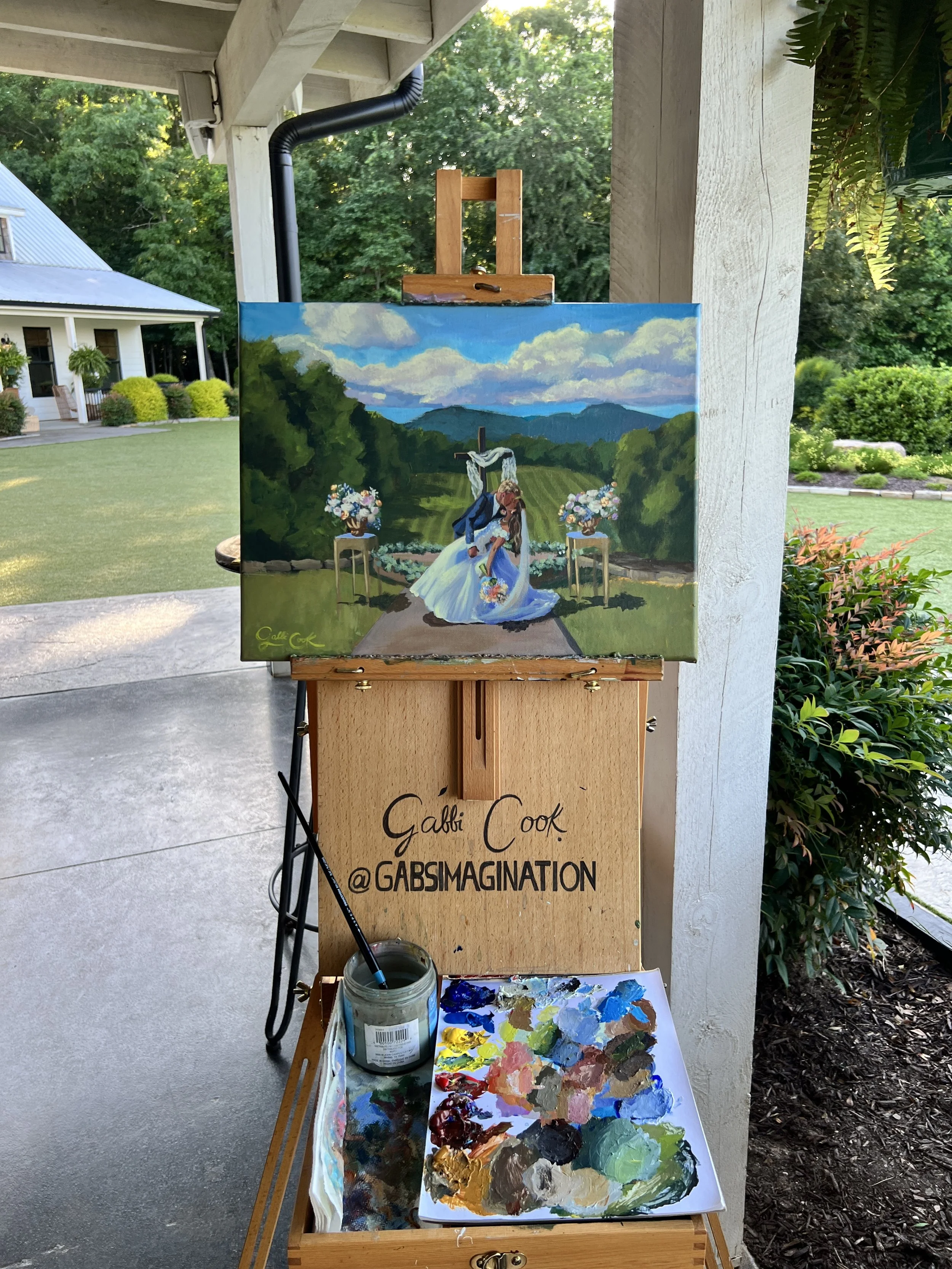 Painting of a wedding scene on an easel, depicting a bride and groom kissing outdoors with mountains in the background. Several flower arrangements are also visible in the painting. In front, a palette with various colors and a jar of paint are on a 
