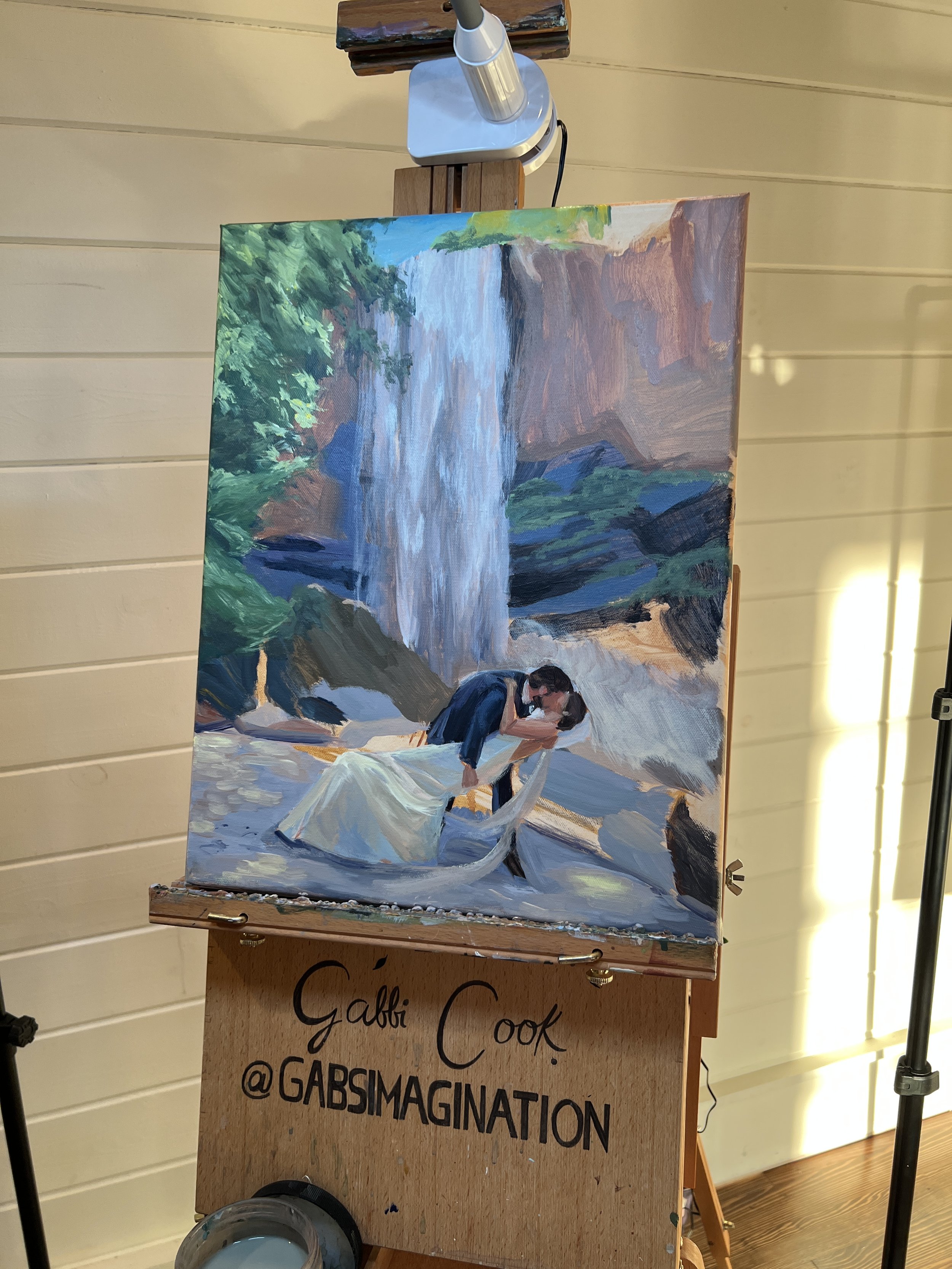 A painting of a couple kissing by a waterfall, with the man in a dark suit and the woman in a white dress, near a rocky terrain and lush greenery.