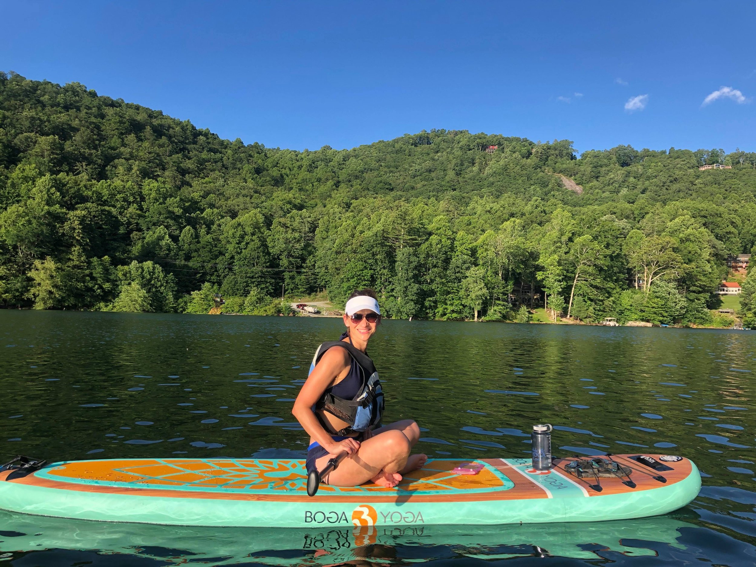 Woman paddleboarding on a lake with green forested hills and a blue sky in the background.