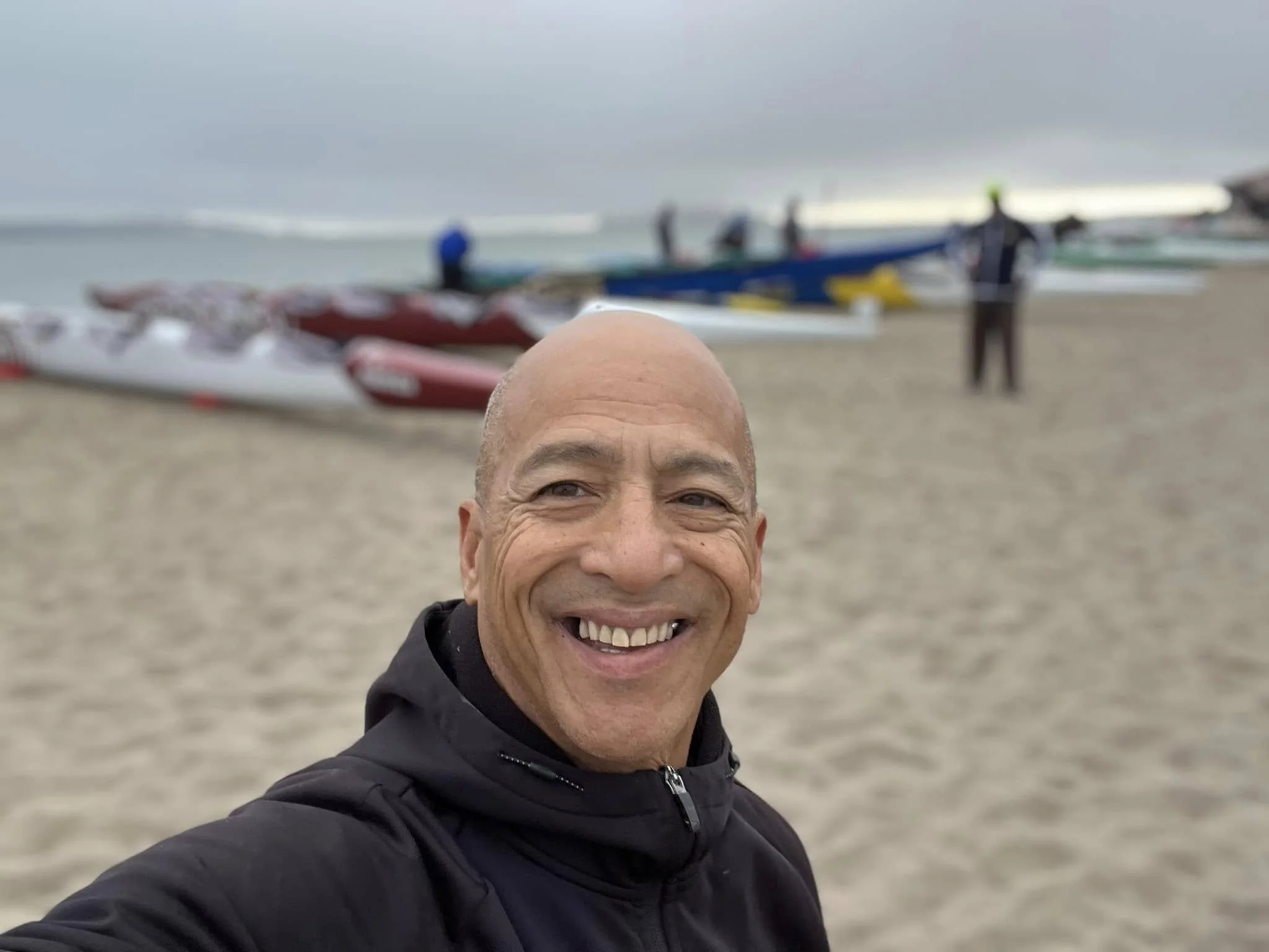 A man taking a selfie on a cloudy beach with kayaks and people in the background.