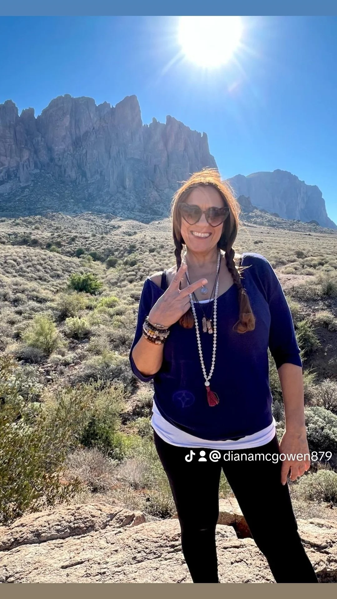 A woman with braided hair and sunglasses smiling outdoors in a desert landscape with mountains and a bright sun in the sky, making a peace sign with her right hand.