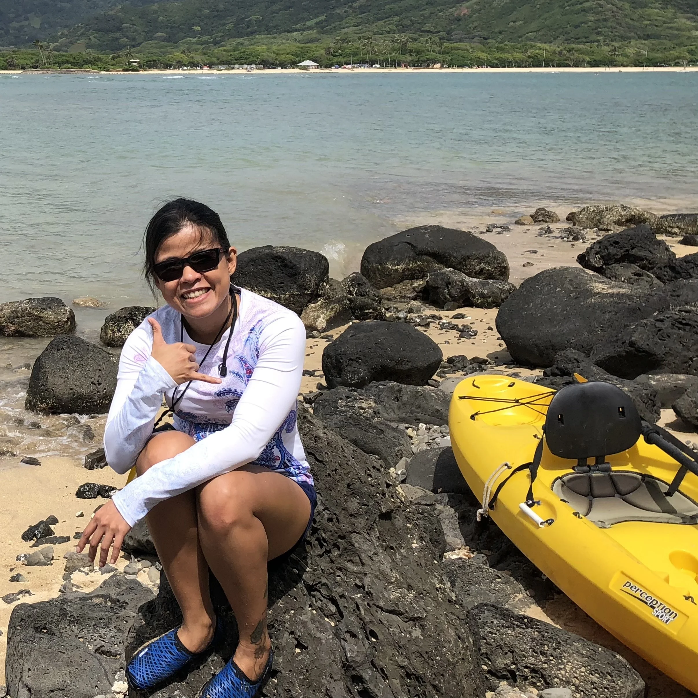 A smiling woman wearing sunglasses, a white long-sleeve shirt, and blue sneakers, sitting on rocks at a beach with a yellow kayak beside her, and the ocean with trees and hills in the background.
