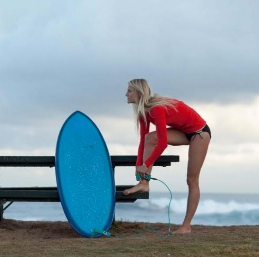 A woman in a red long-sleeve shirt and black shorts is preparing to paddleboard at the beach. She is tying her paddleboard to a bench with a blue leash, with the ocean and cloudy sky in the background.