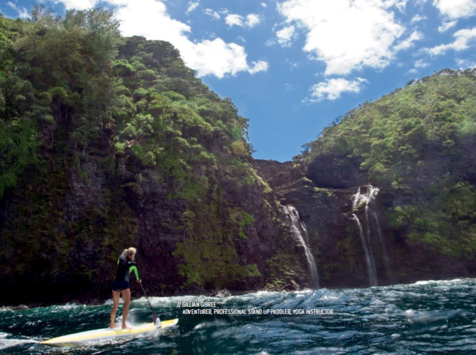 A person paddleboarding on the ocean near lush green cliffs with waterfalls, under a partly cloudy sky.