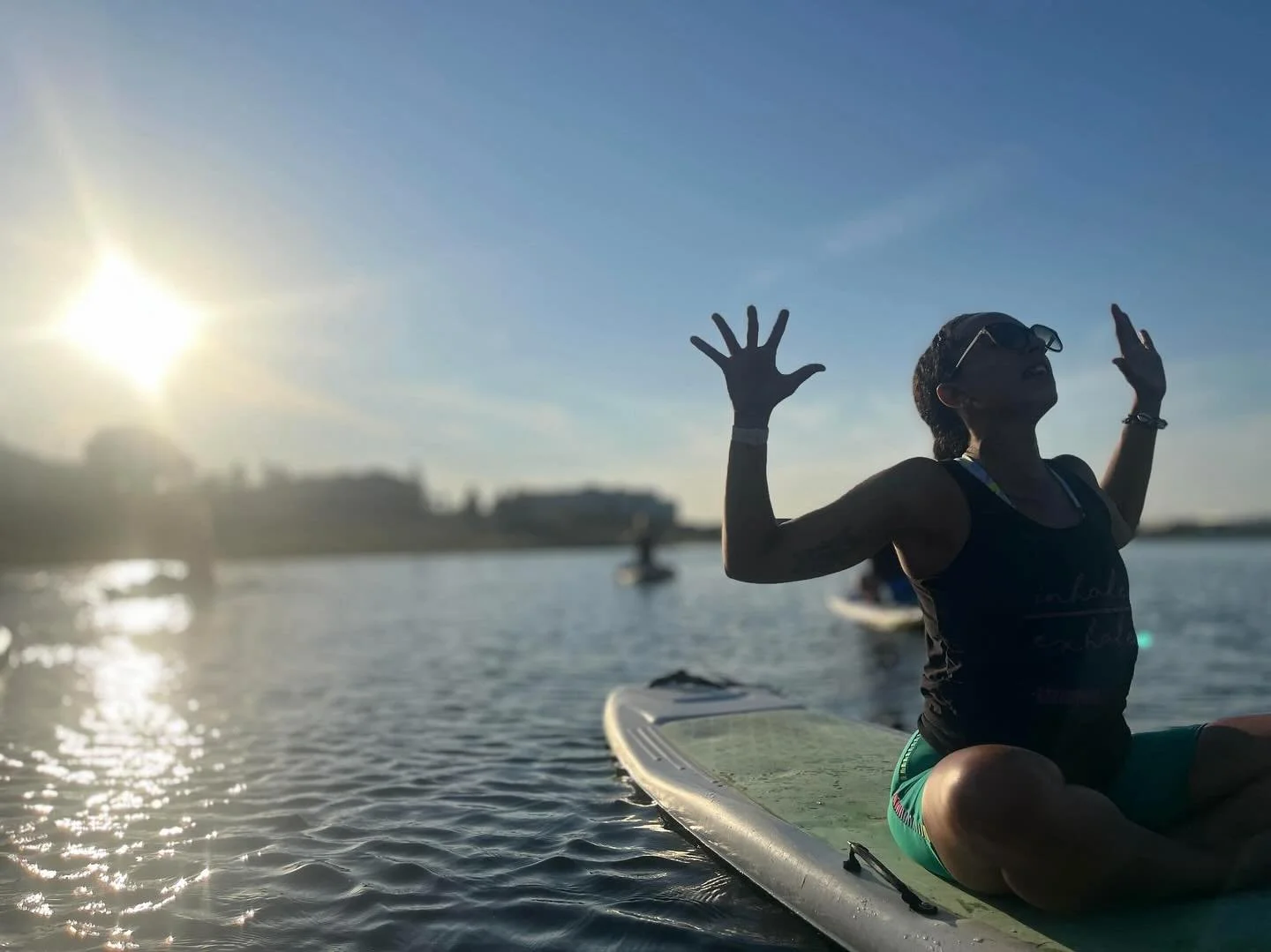 Person sitting on paddleboard on water near sunset, raising hands and facing the sky.