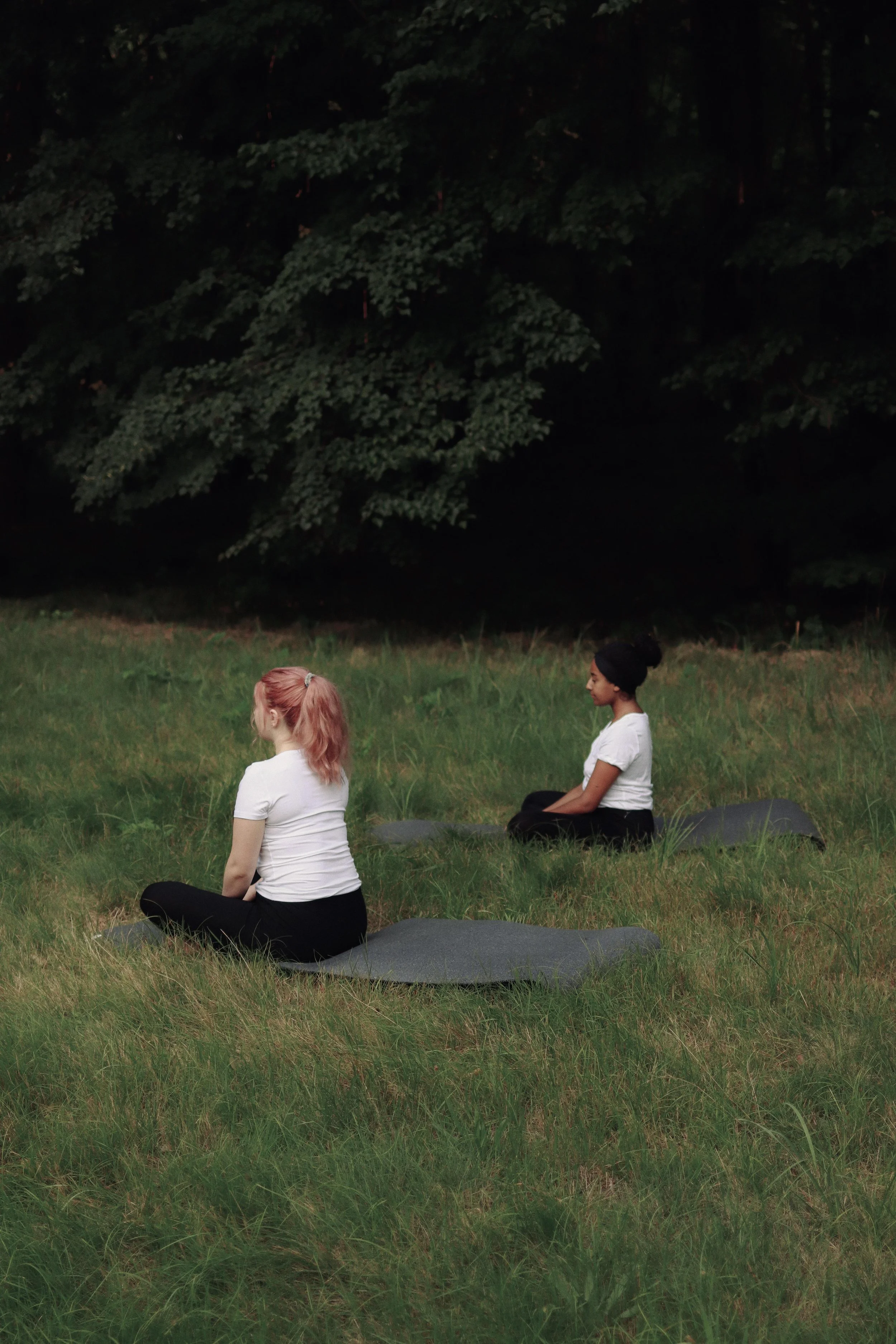 Zwei Frauen sitzen auf Yogamatten auf einer Wiese am Wald und atmen ruhig.