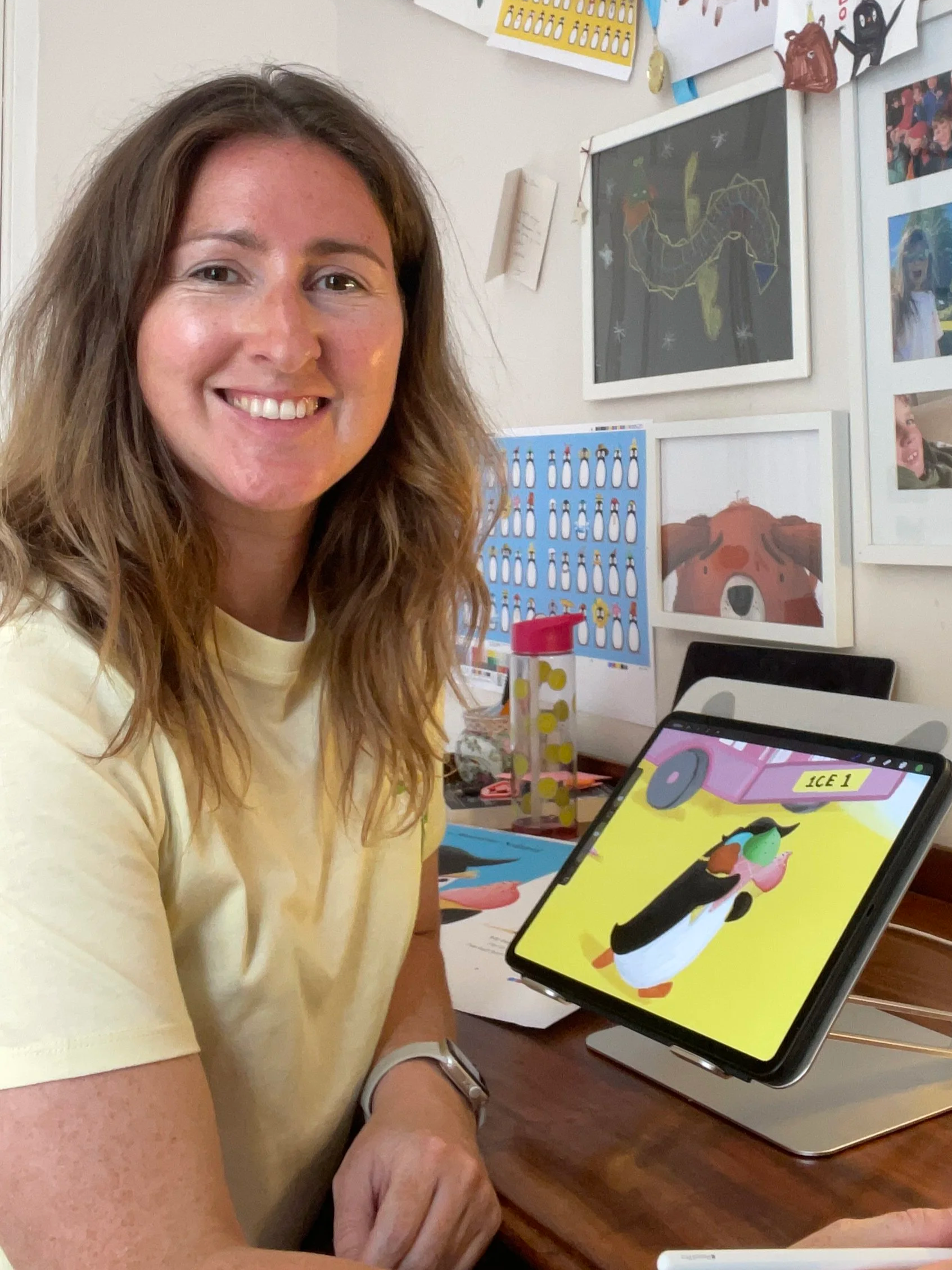 Smiling woman seated at a desk beside a tablet displaying a colorful penguin illustration, with children’s artwork and photos on the wall behind her.