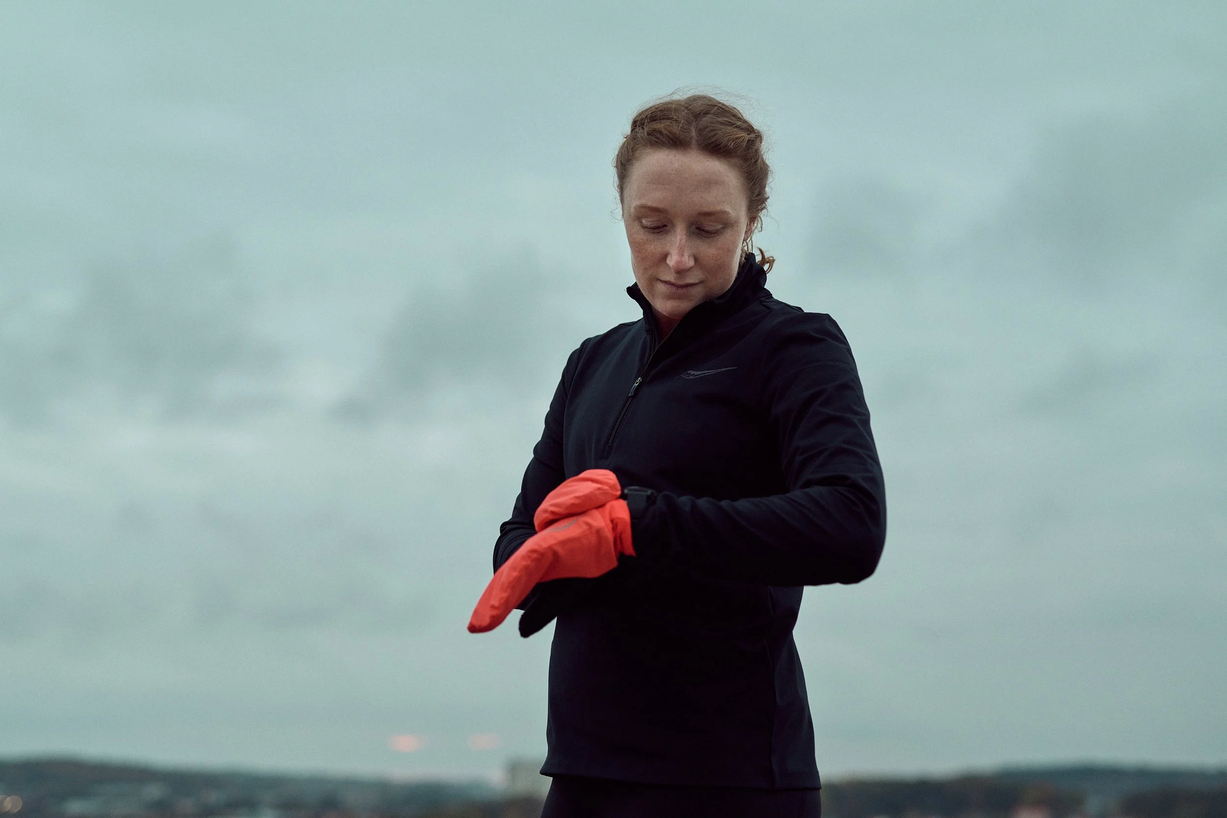 A woman with reddish hair looking down, wearing a black jacket and orange gloves, outdoors on a cloudy day.