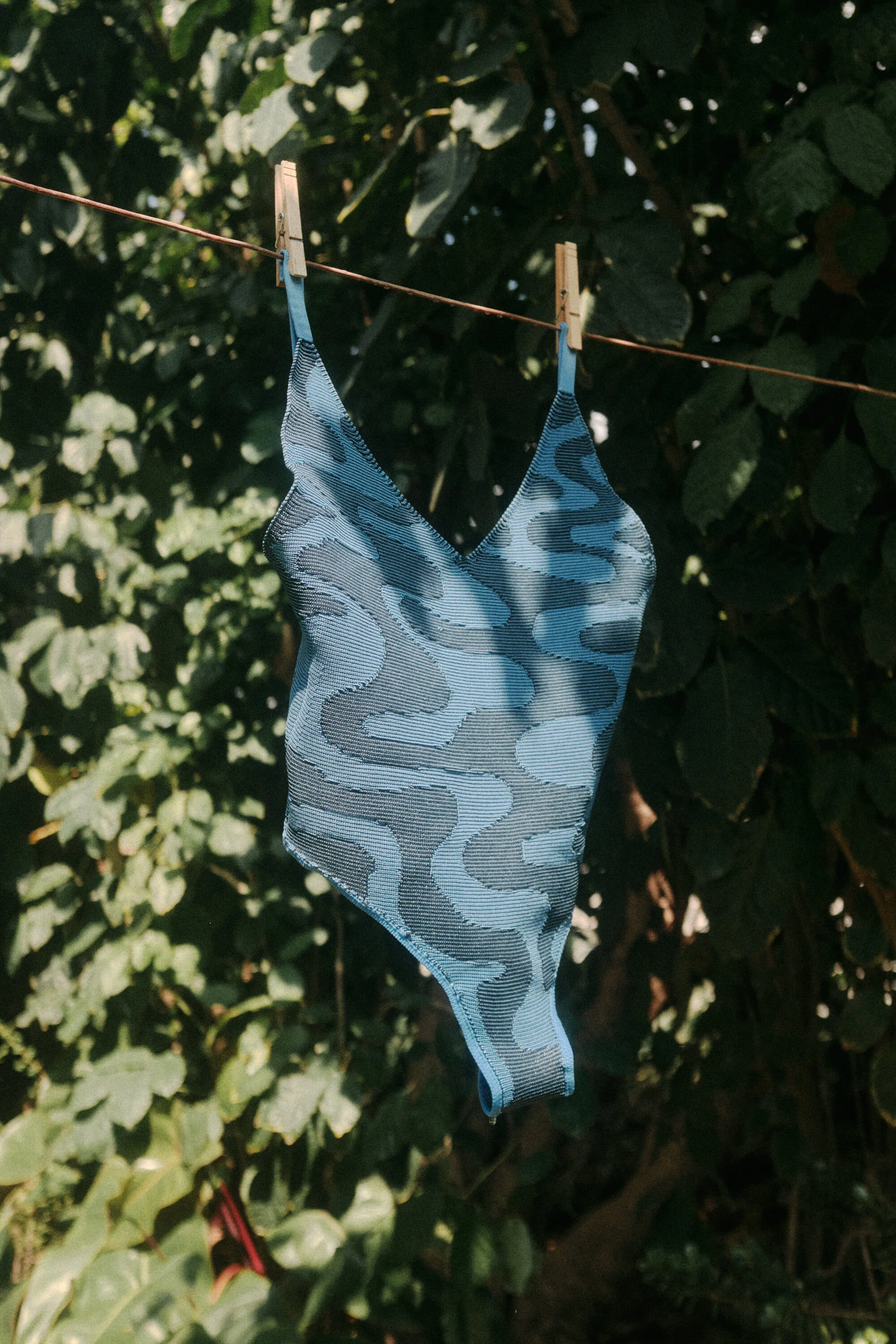 A blue and black patterned face mask hanging on a pink clothesline with two wooden clothespins against a background of green foliage.