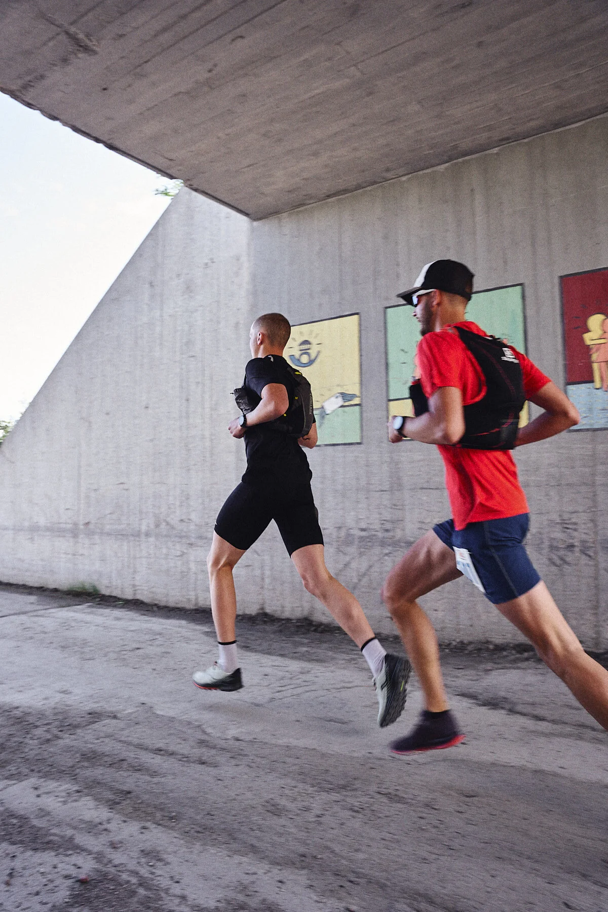 Two men running outdoors near a concrete wall with colorful abstract art. One wears a black shirt and shorts, the other a red shirt and dark shorts. They carry hydration packs and wear running shoes.