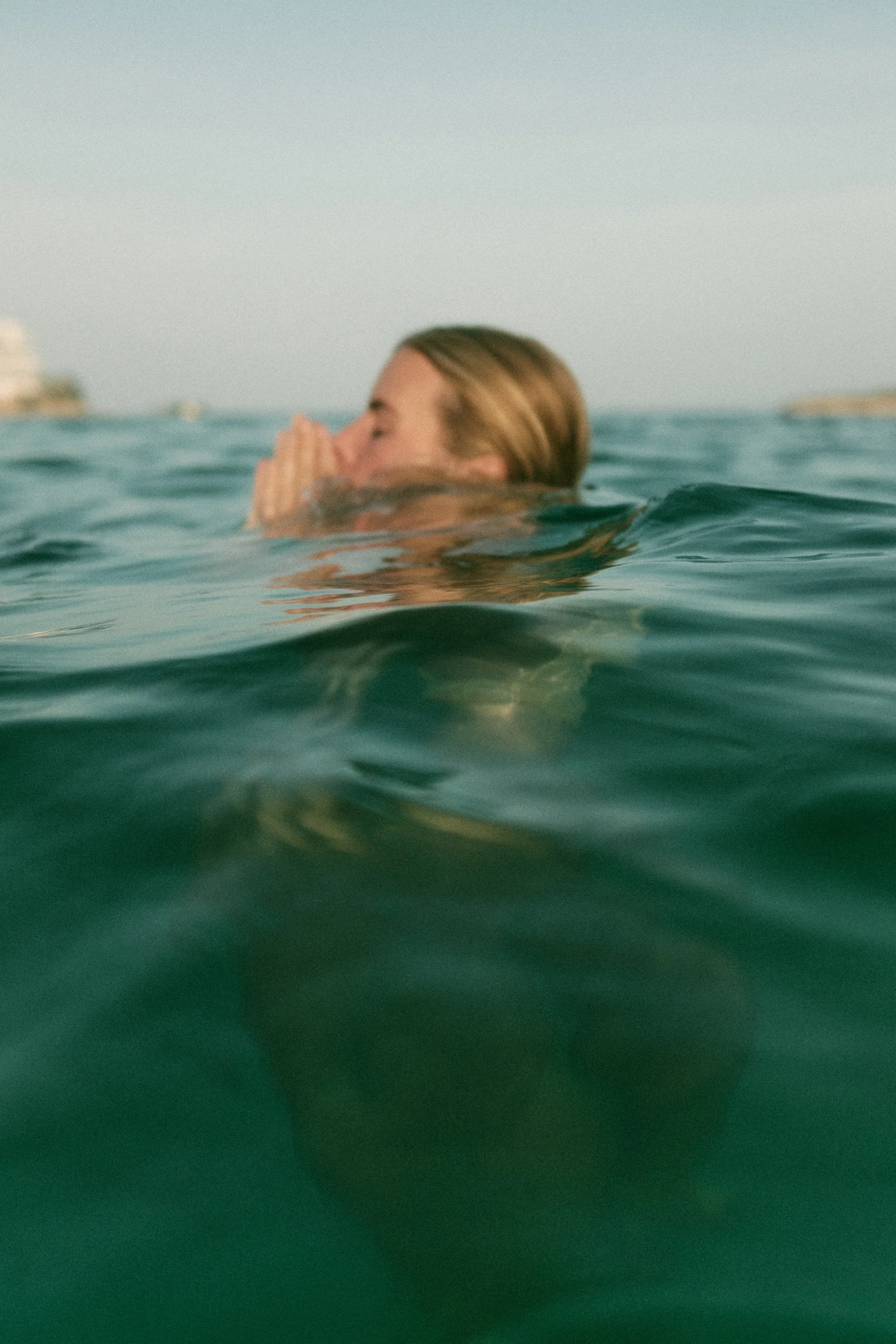 A woman swimming in the ocean, with her face and hands lifted above the water, eyes closed and praying or meditating.