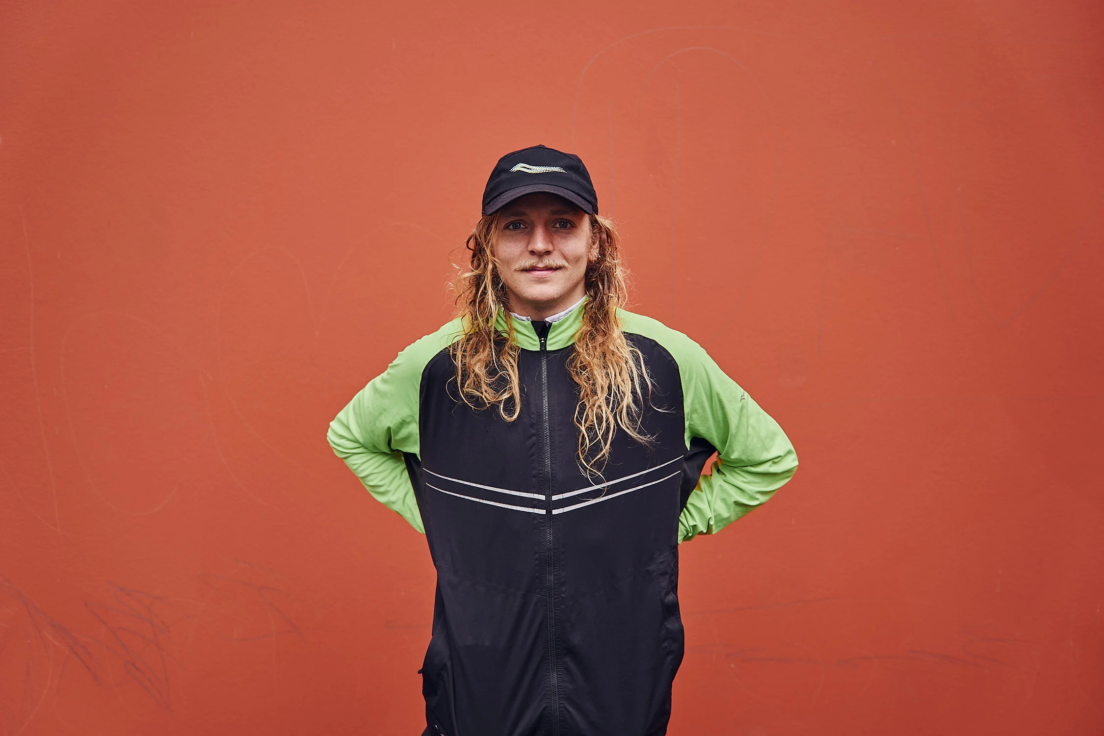 A person with long curly hair, wearing a black baseball cap and a black and neon green athletic jacket, standing with hands on hips in front of an orange wall.