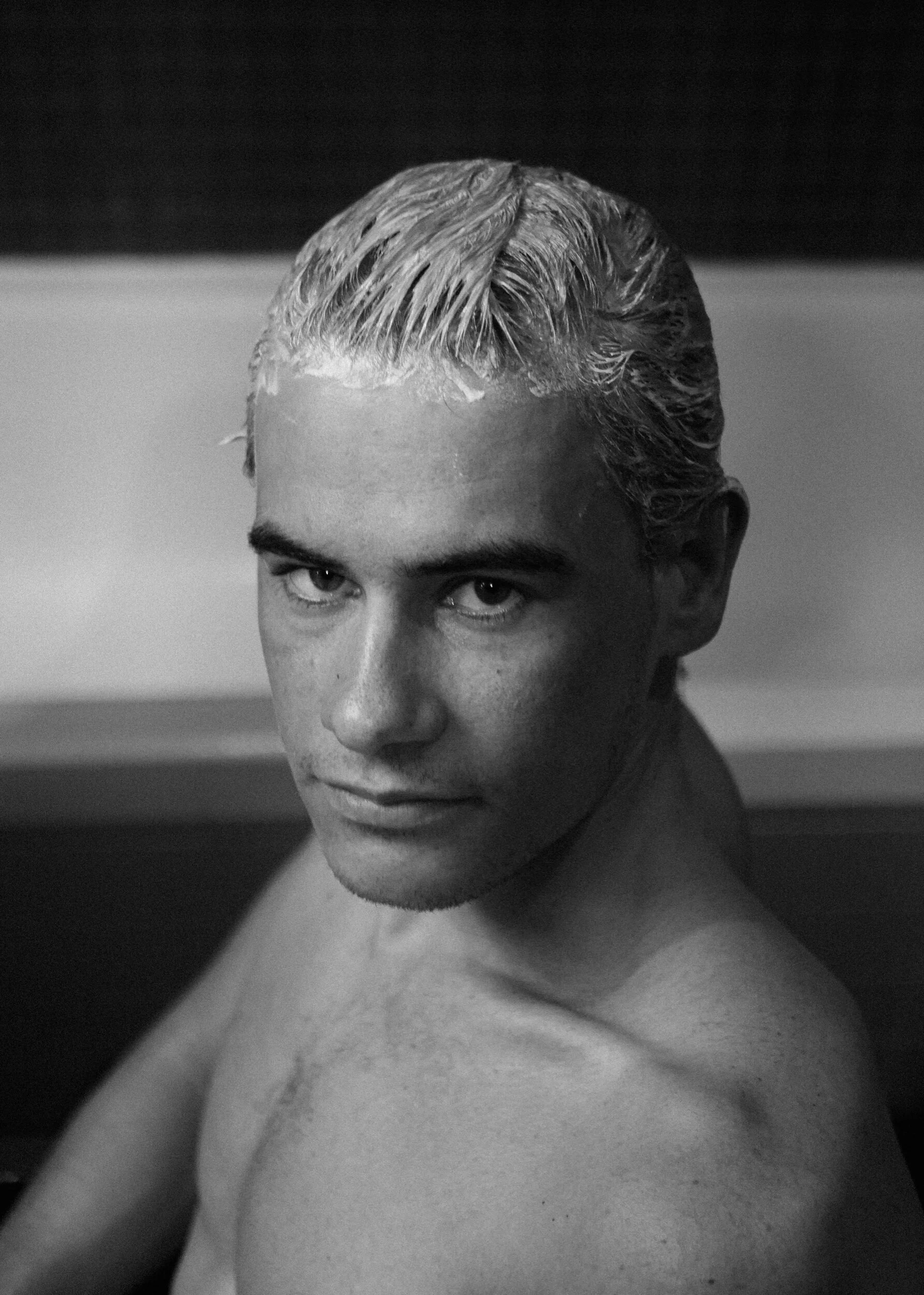 Black and white portrait of a young man with short, wet hair, looking directly at the camera with a neutral expression, top of his head to his shoulders visible.