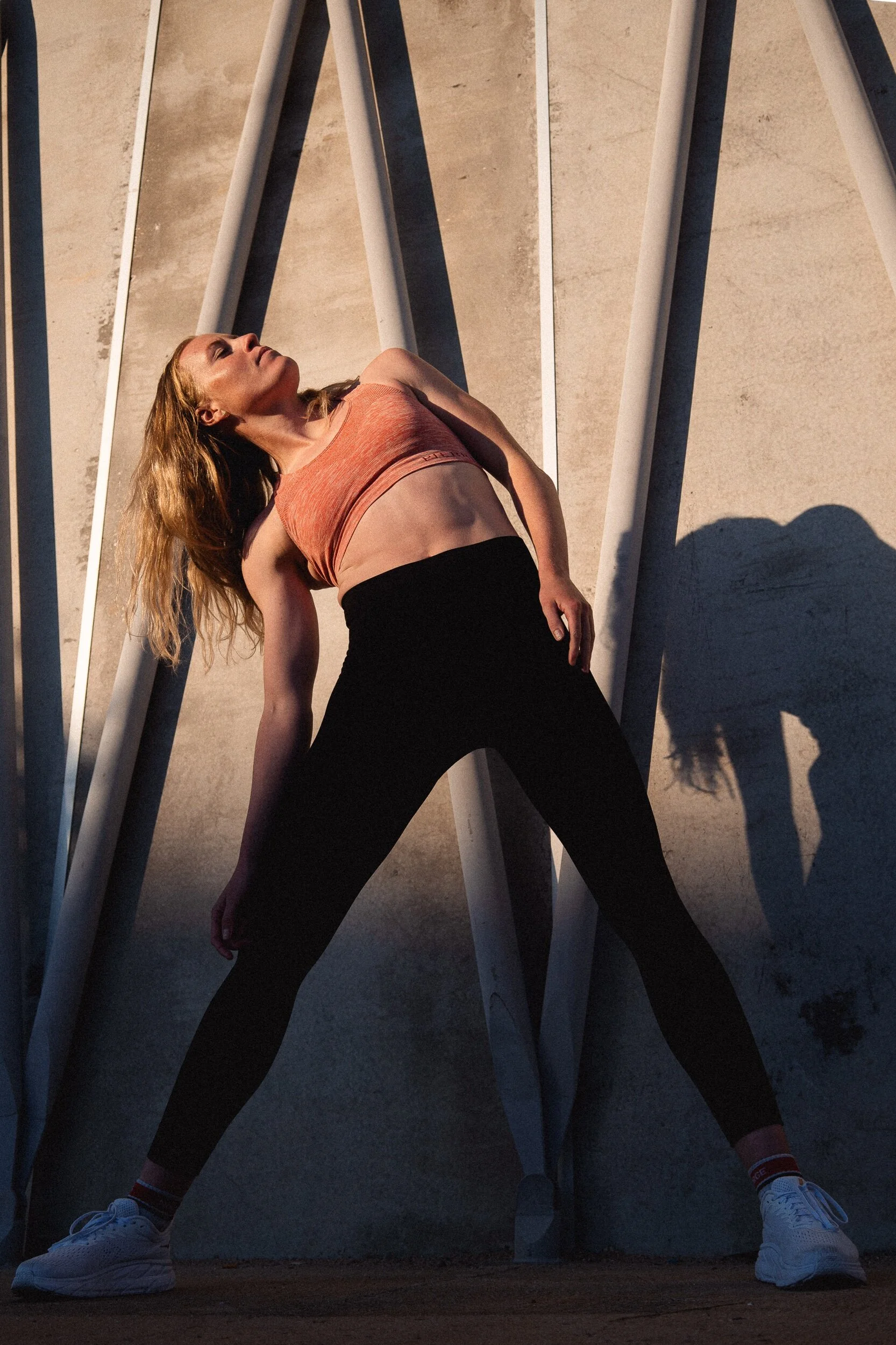 A woman wearing a peach sports bra, black leggings, and white sneakers is standing outdoors, leaning back with her hands on her thighs, surrounded by metal bars casting shadows on the concrete ground.