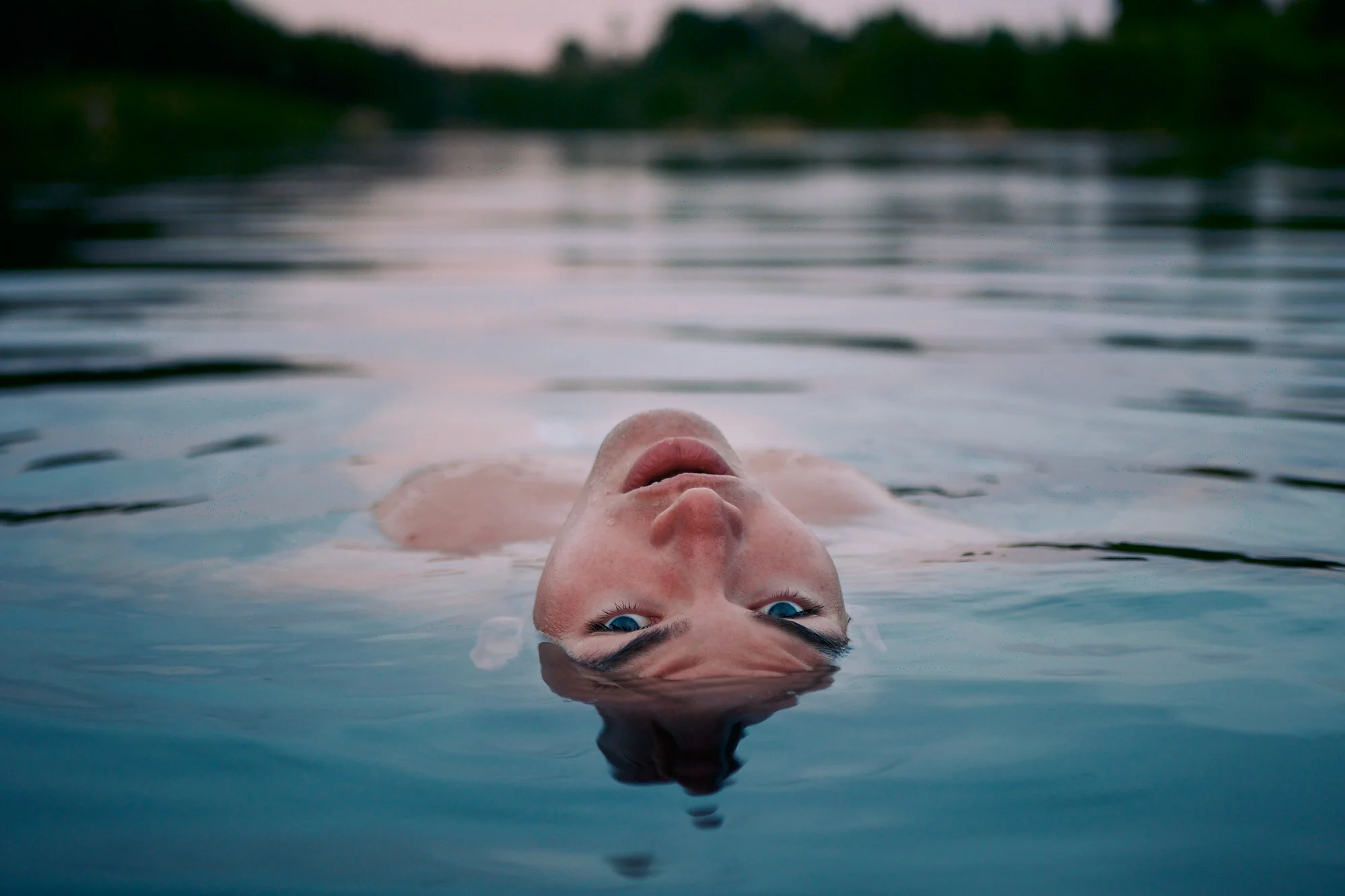 A child with blue eyes floating face-up in a body of water, with the water's surface reflecting the sky and a blurred tree line in the background.