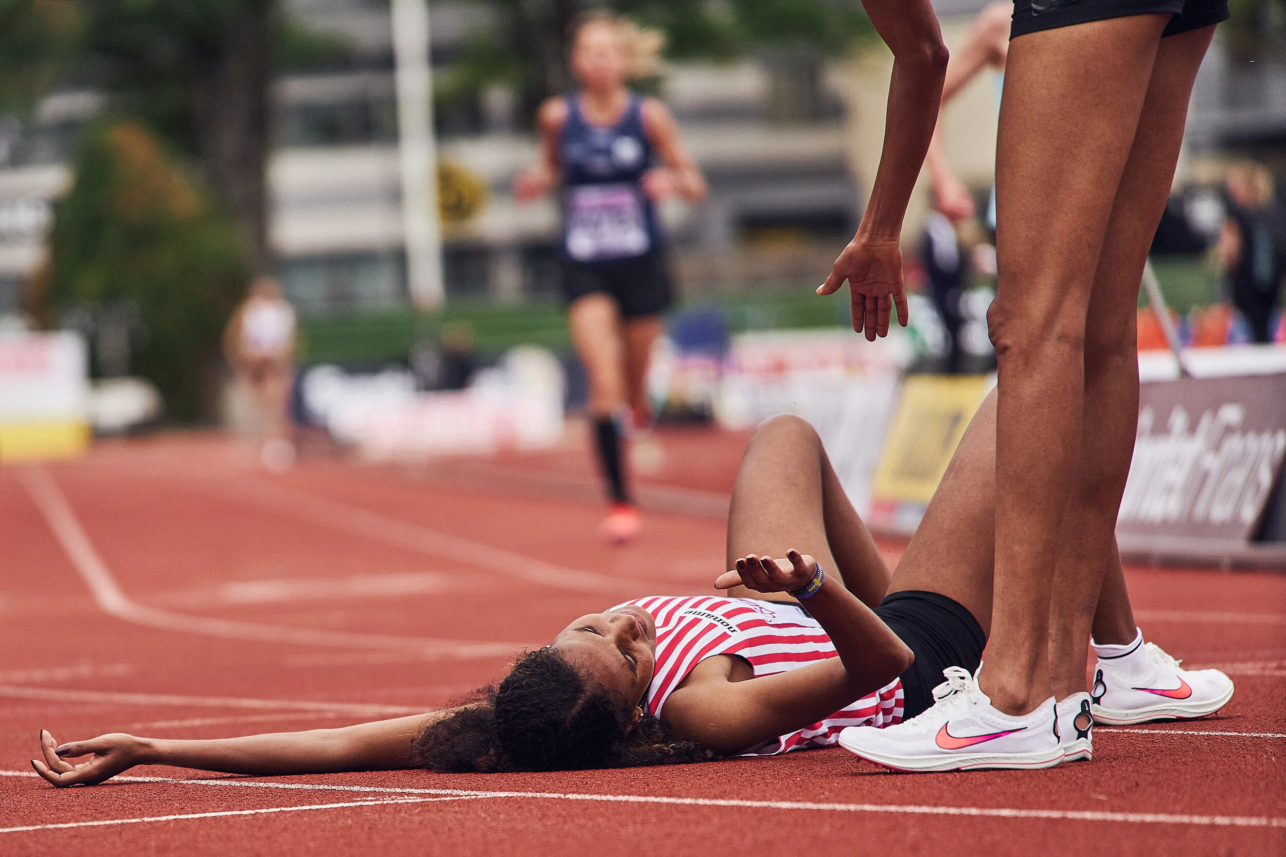 An athlete lying on the track after falling during a race, with another athlete helping her up. The scene takes place on a red running track with a blurred background of other runners and spectators.
