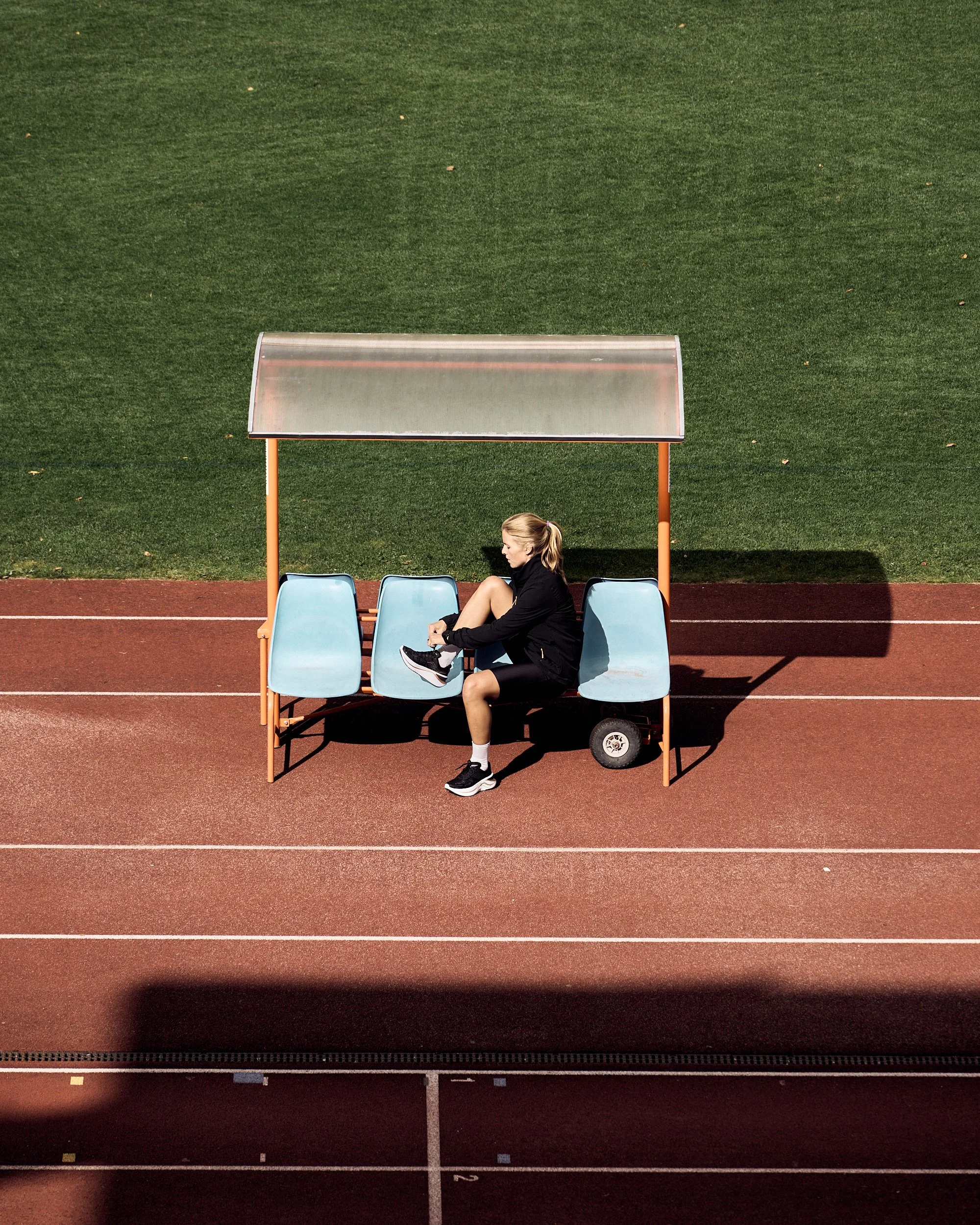 A young woman in black athletic clothing is tying her shoe while seated on a light blue bench under a small covered shelter on a red running track. Behind her is a large green grassy area.